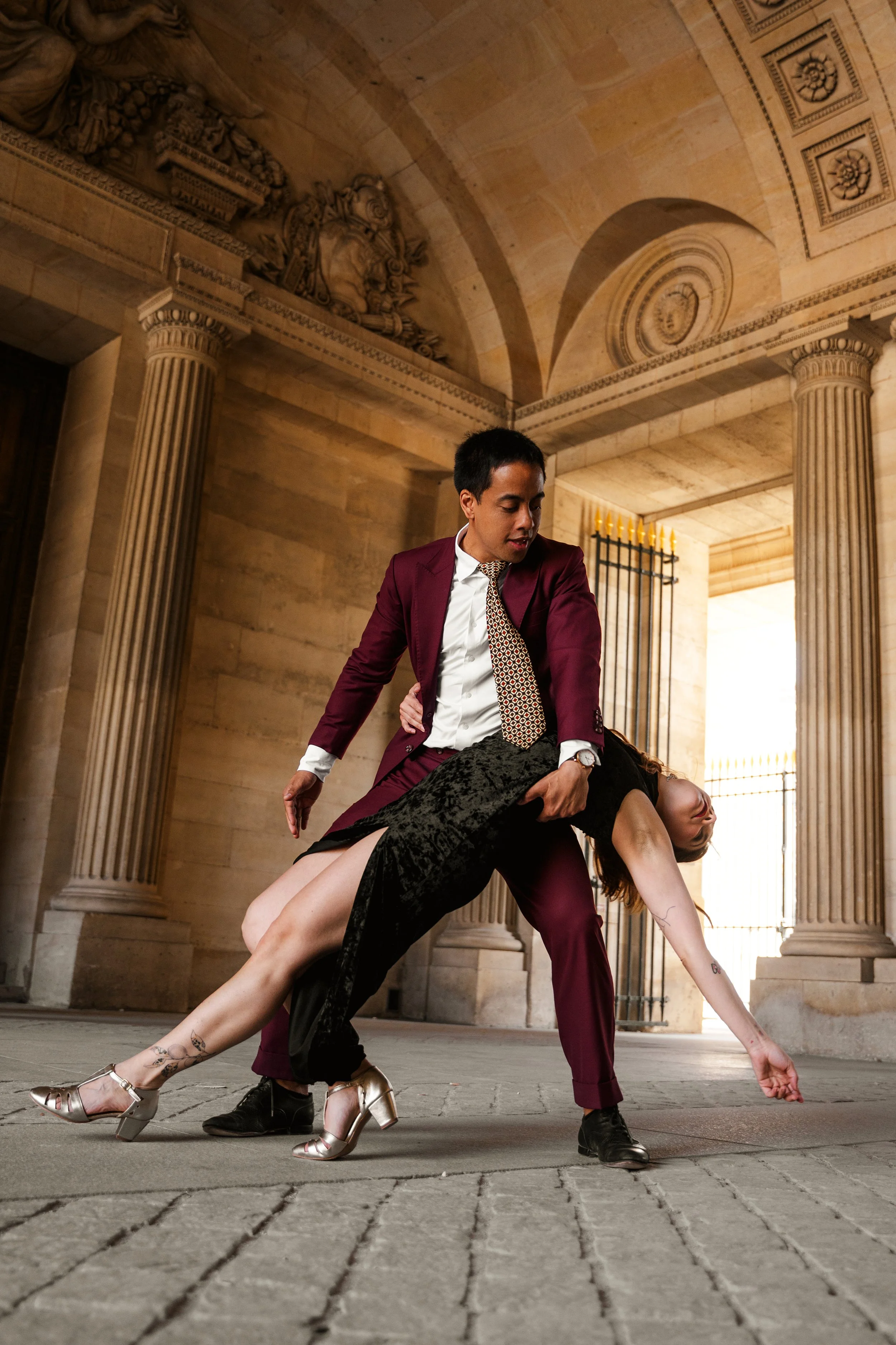 Un homme en costume bordeaux et une femme en robe noire en velour fendue, font un tombé de danse, dans une architecture ancienne avec des colonnes et un plafond voûté.