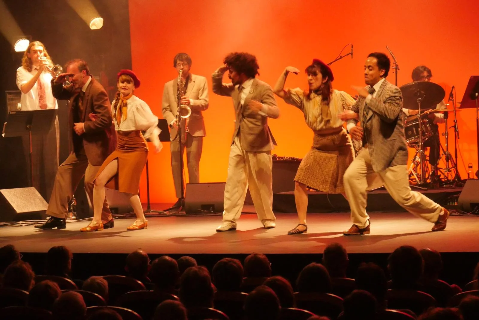 Une scène de spectacle avec plusieurs danseurs en costumes années 1940 sur scène, accompagnés par un groupe de musiciens de jazz.