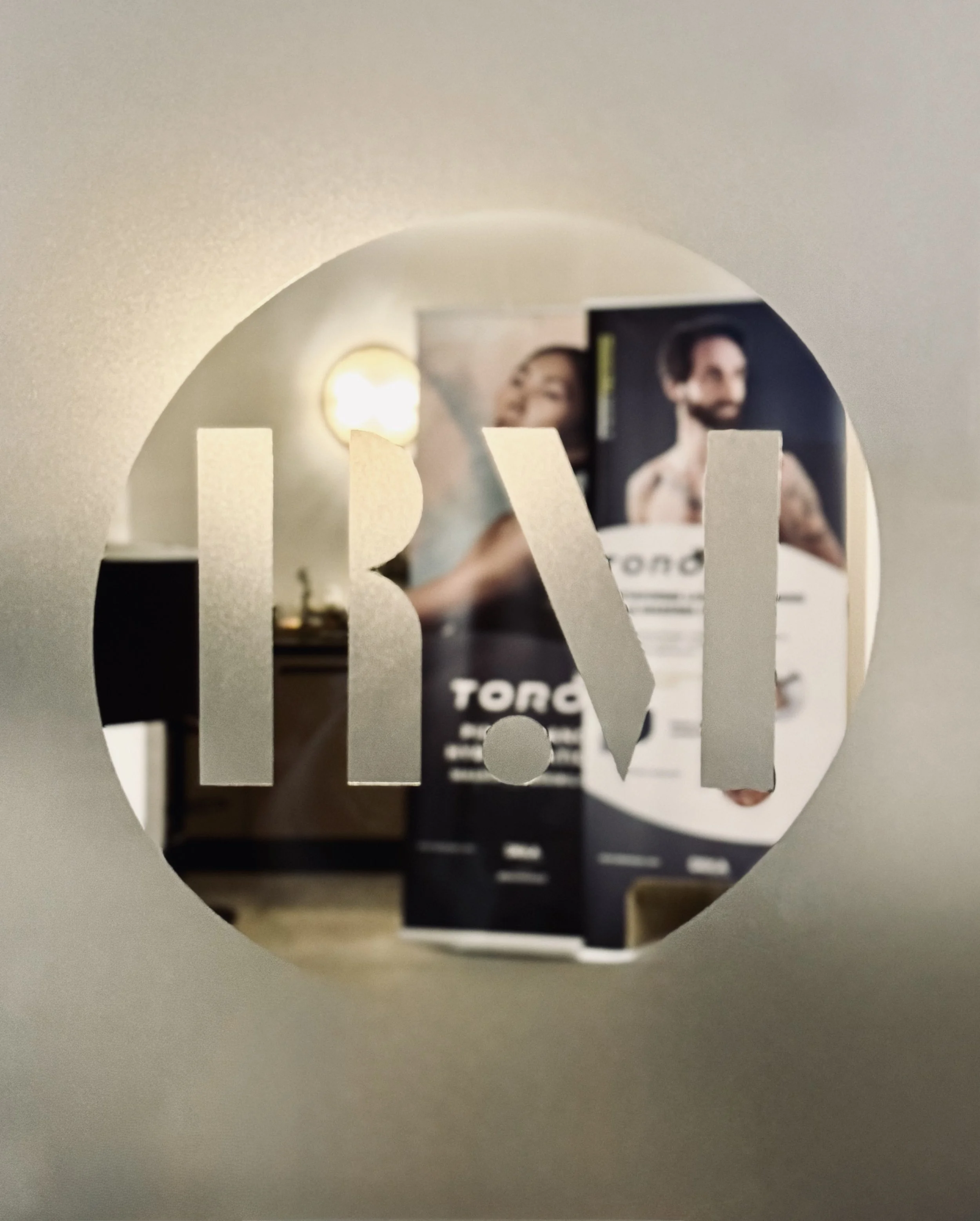 Vista attraverso un cerchio con il logo NBR in metallo, che rivela una parete con manifesti di presentazione di prodotti per parrucchieri, incluso un uomo con tatuaggi e una donna con capelli ricci.