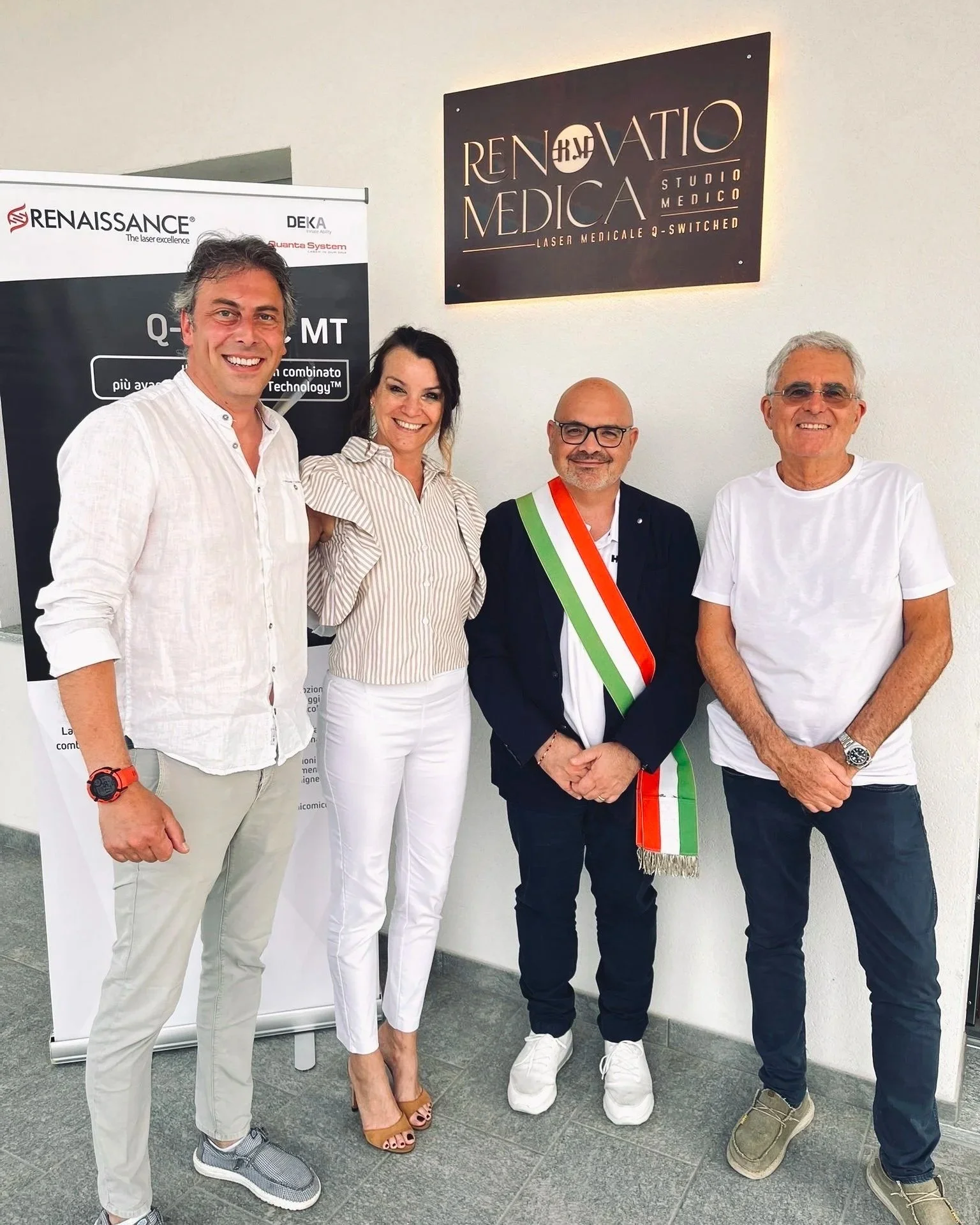 Quattro persone sorridenti davanti a un'insegna che recita "Renuvatio Medica" e altri segni pubblicitari.