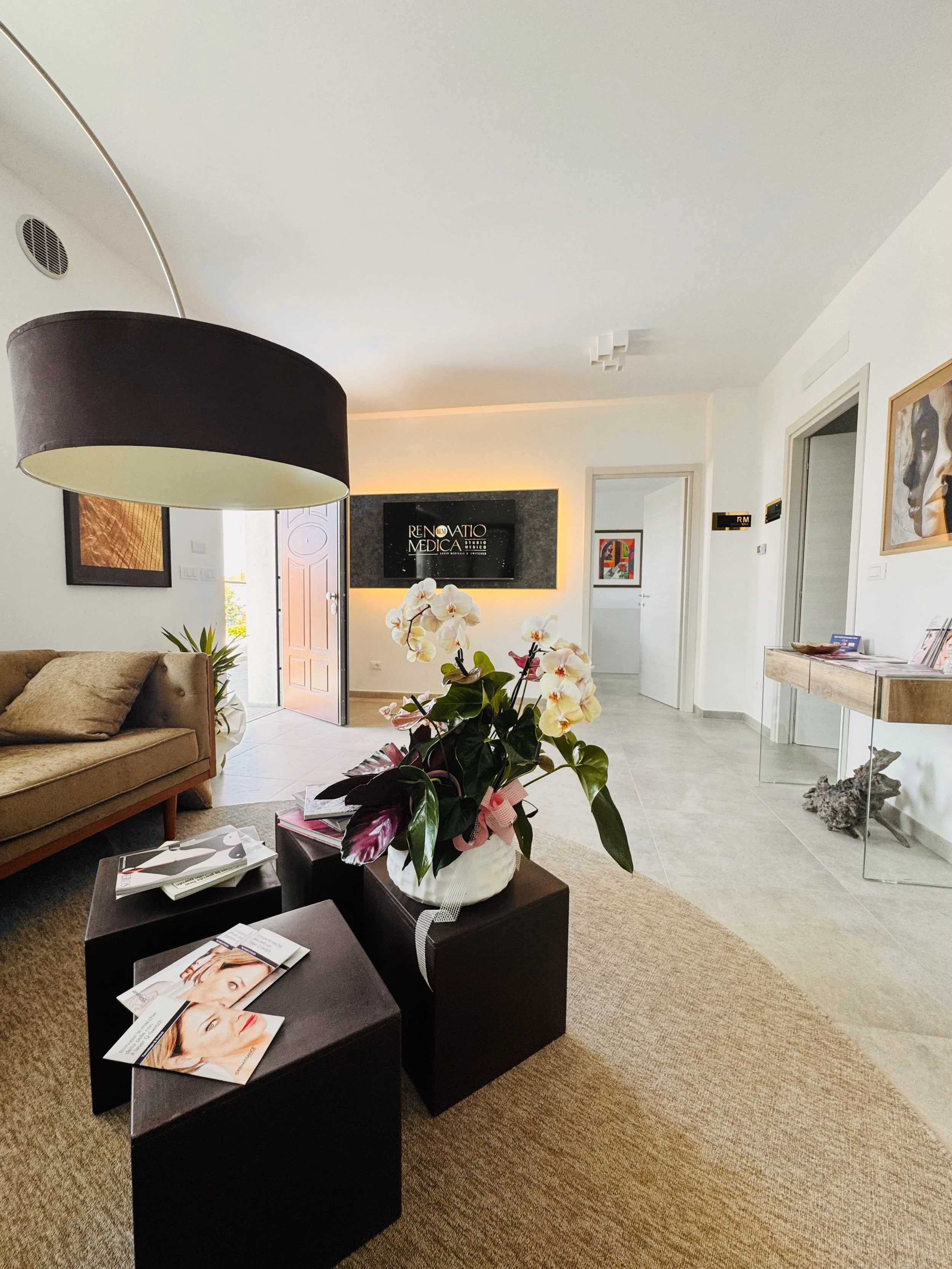 Reception di uno studio medico con sedia beige, poltrona in tessuto, tavolini neri con riviste, fiori bianchi in vaso, pareti bianche con quadri, porta aperta e schermo con scritta "Rinnovato Medica".