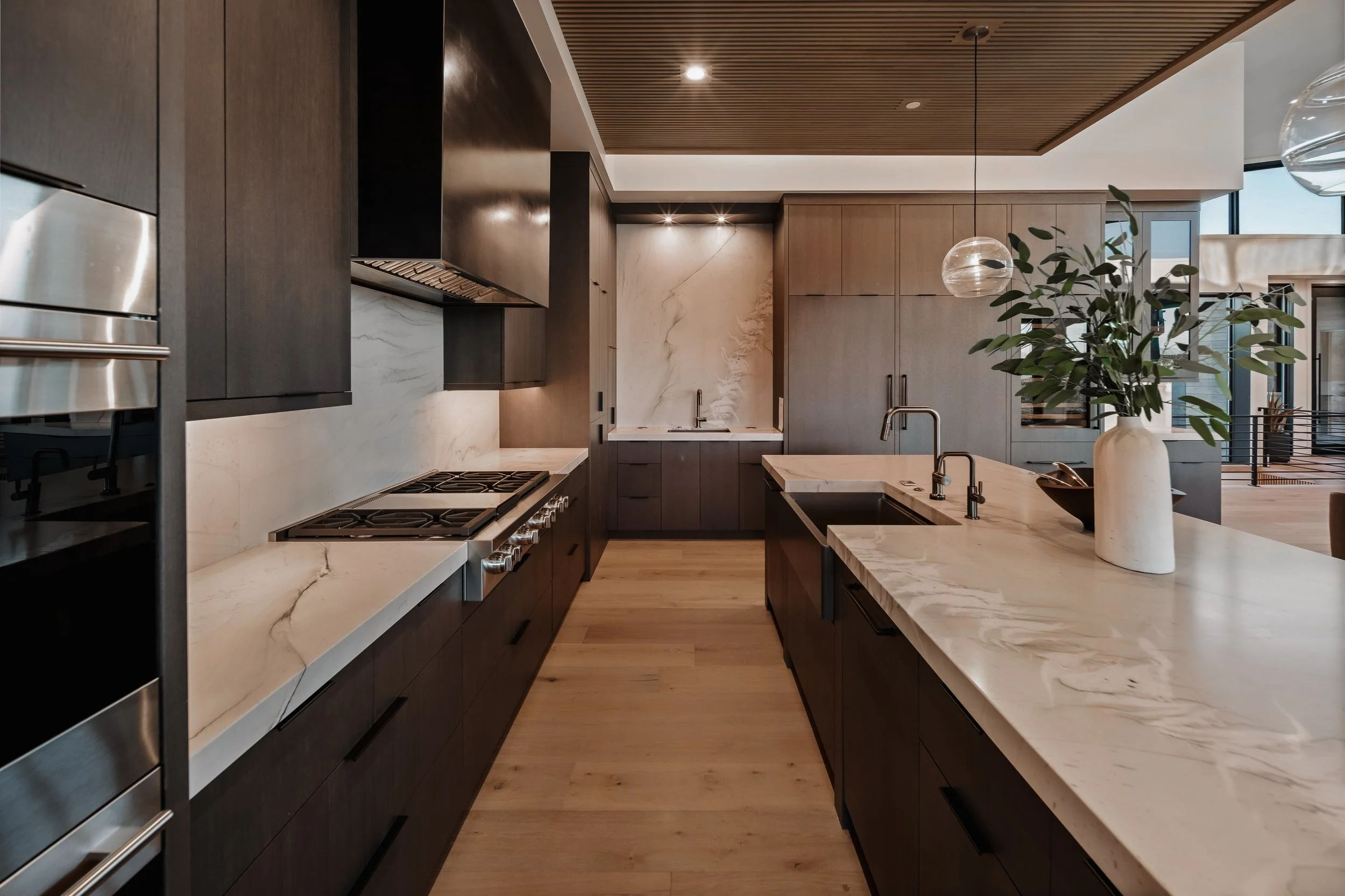 Modern kitchen with dark wood cabinets, white marble countertops, a built-in oven, gas stove, and a vase with green leaves on the island.