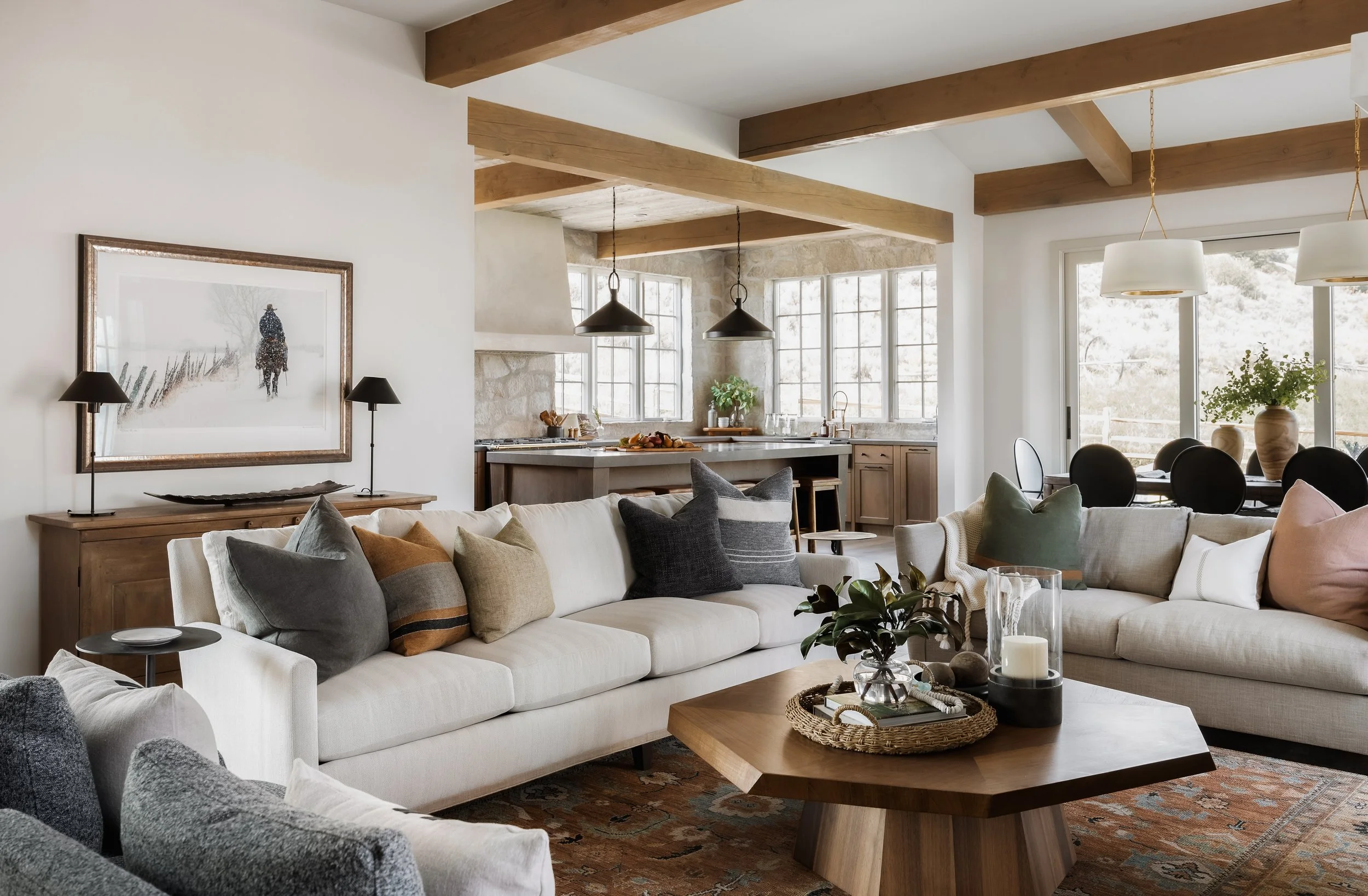 Living room with cream sofas, decorative pillows, wooden coffee table, and a sideboard with lamps. An open kitchen with large windows, black pendant lights, and beige cabinetry is visible in the background. Dining area with a table, chairs, and potte