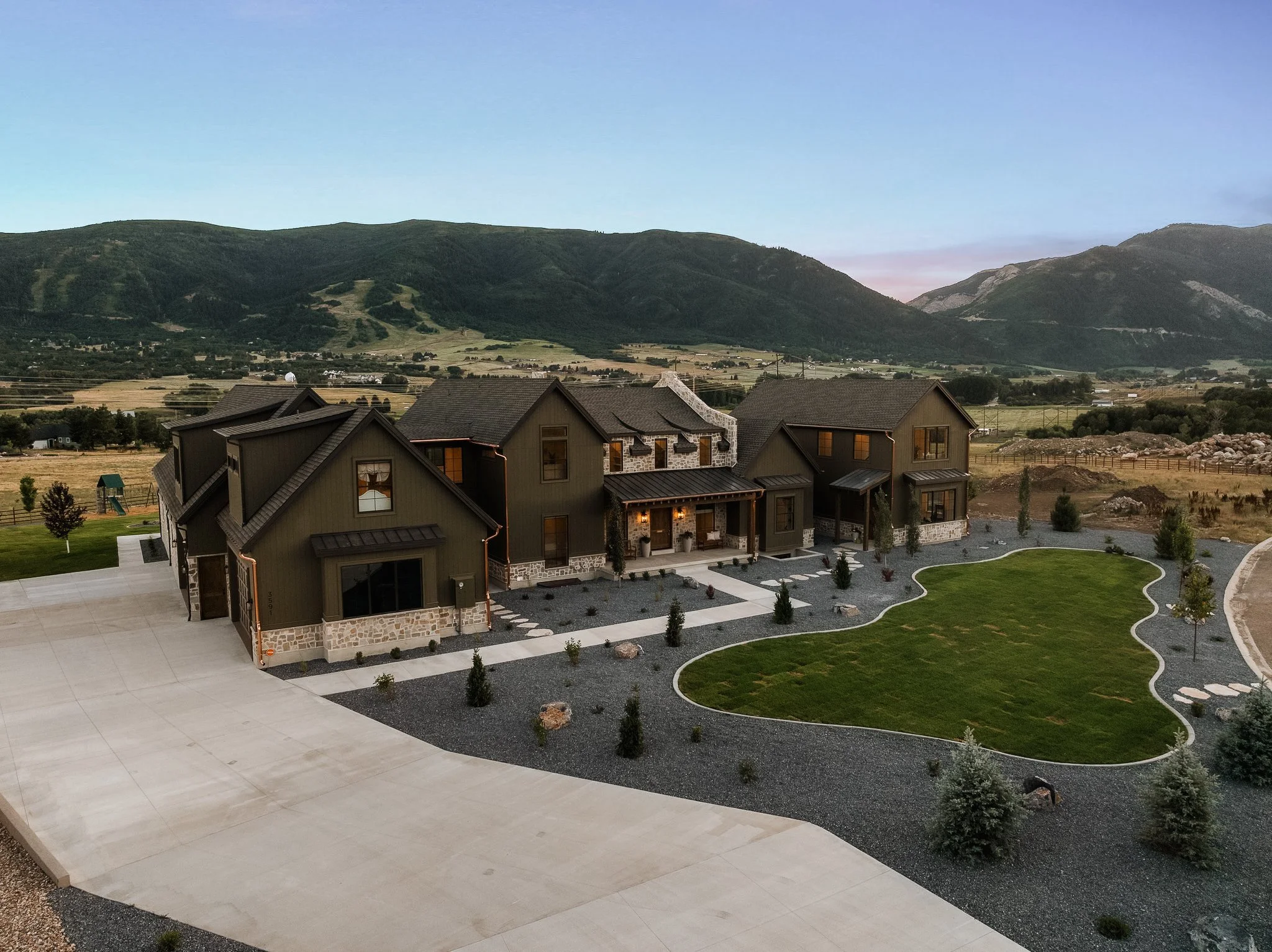 A large modern house with dark exterior walls, stone accents, and a metal roof, surrounded by landscaped yard with a curved grass patch, small trees, and gravel pathways, set against a backdrop of green mountains and open countryside.