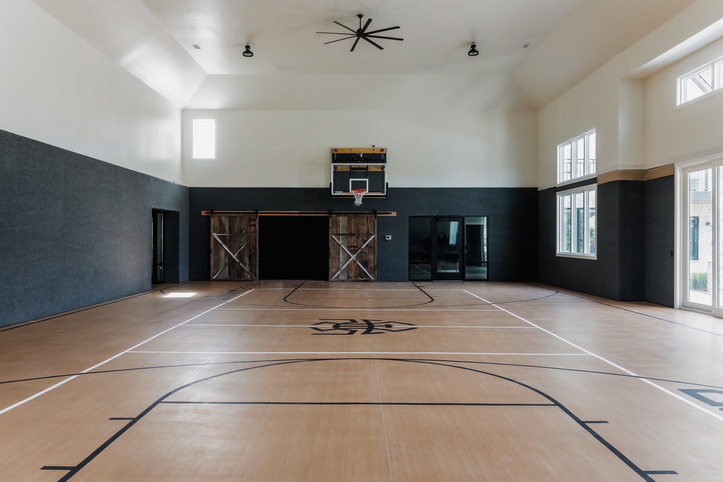 Empty indoor basketball court with wooden flooring, black and white lines, a basketball hoop on the back wall, large windows on the right side, and ceiling fans.