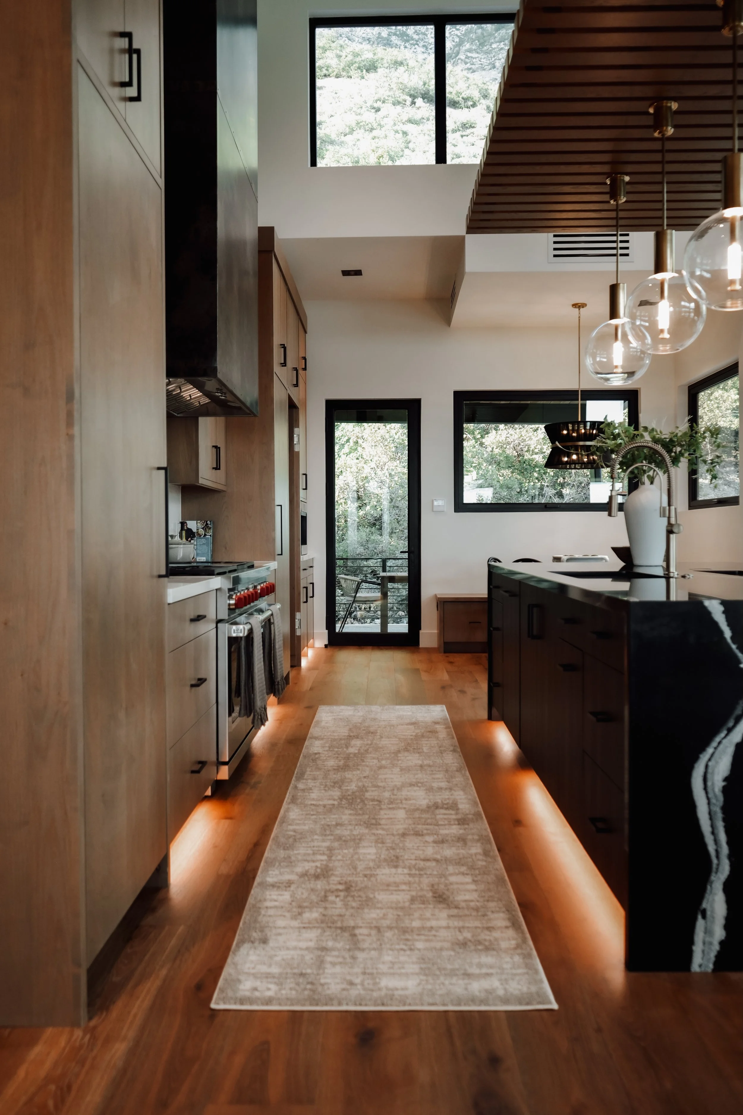 Modern kitchen with wood cabinets, black countertops, pendant lights, large windows, and a sliding glass door leading outside.