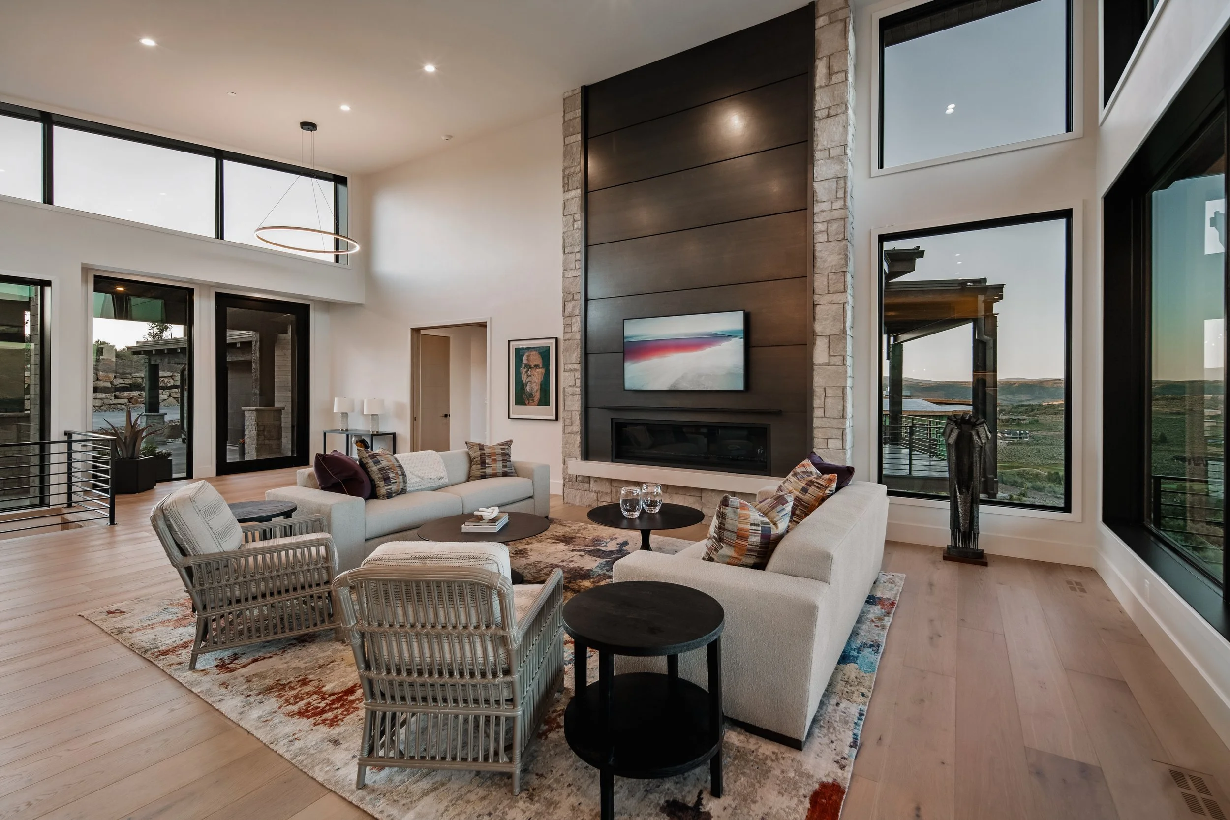 Modern living room with large windows, a stone and wood fireplace, white sofas, armchairs, and a patterned area rug, overlooking a scenic outdoor landscape.
