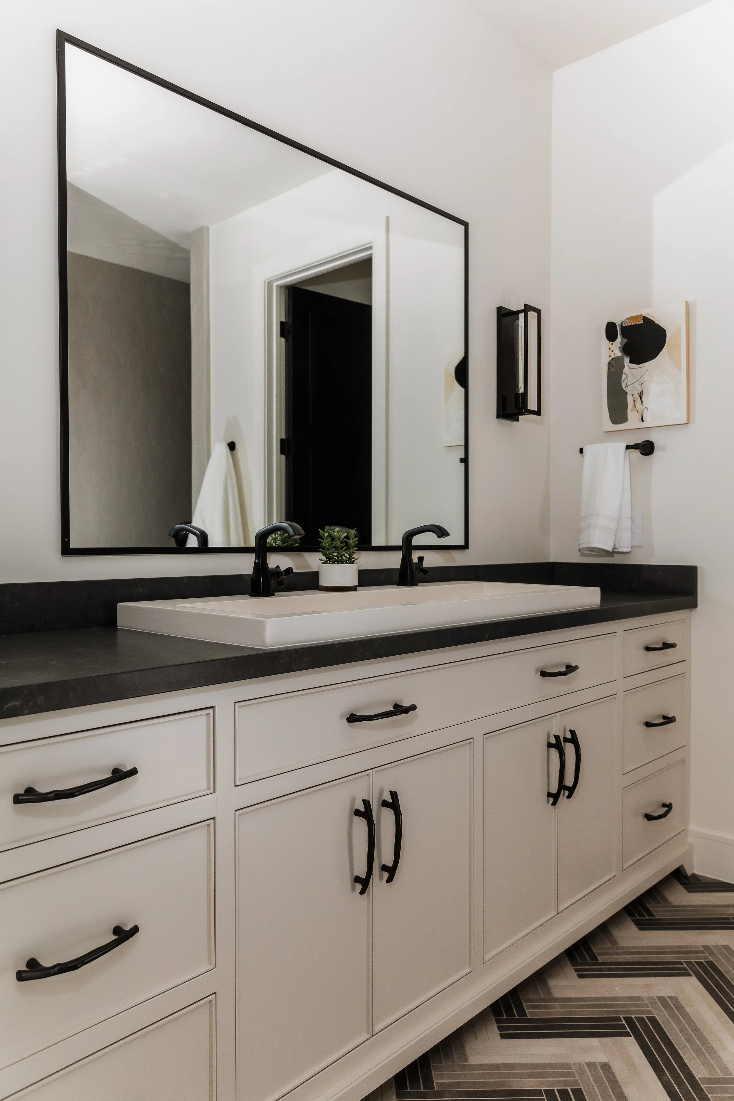 Modern bathroom vanity with white cabinets, black handles, a black countertop, and a white sink with black faucets. A large rectangular mirror is above the sink, reflecting part of the bathroom. There is a small potted plant on the counter, a black w