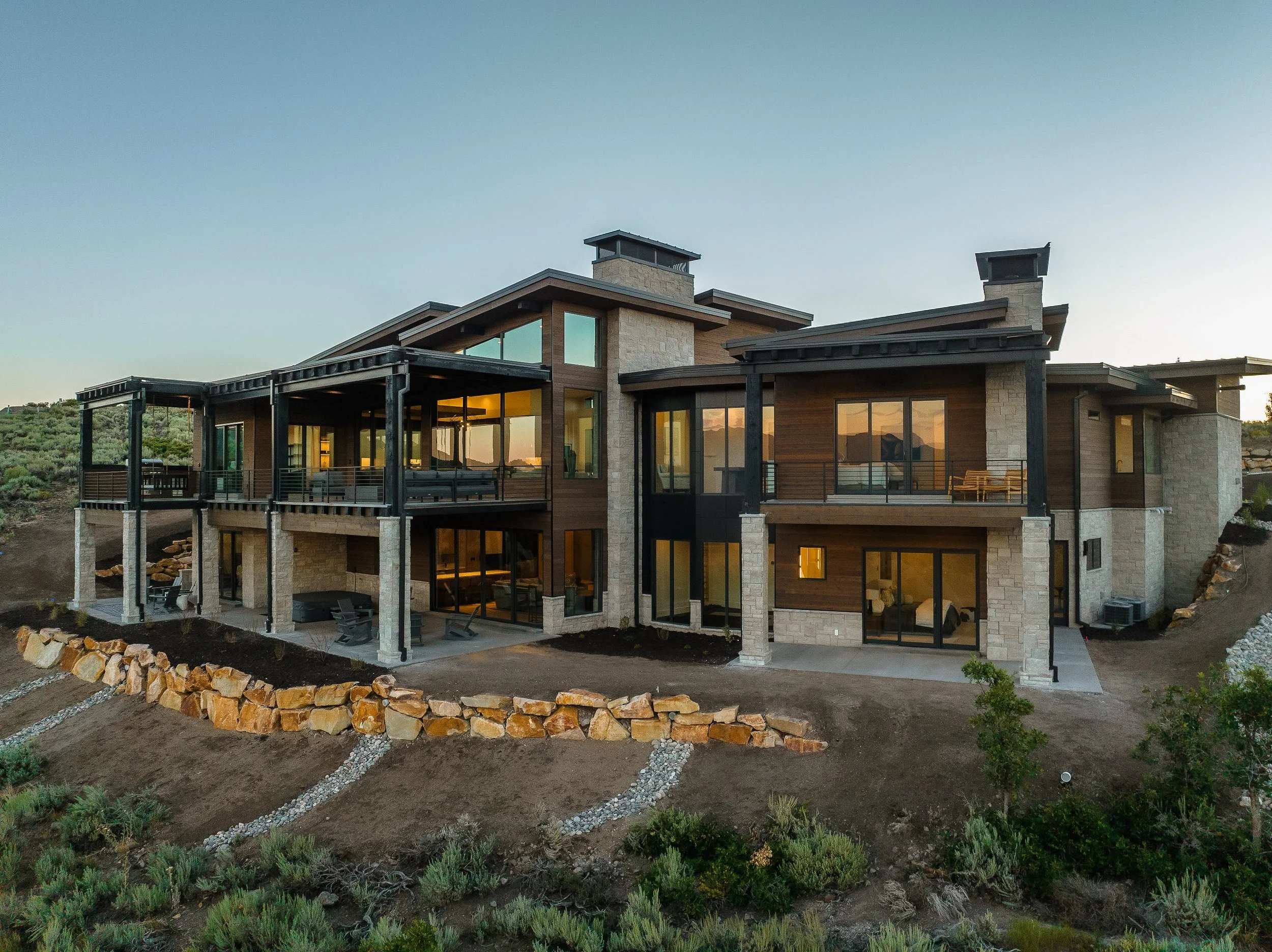 Modern multi-story house on a hillside with large windows, stone and wood exterior, and outdoor living spaces. The house is surrounded by desert vegetation and a stone-lined pathway.