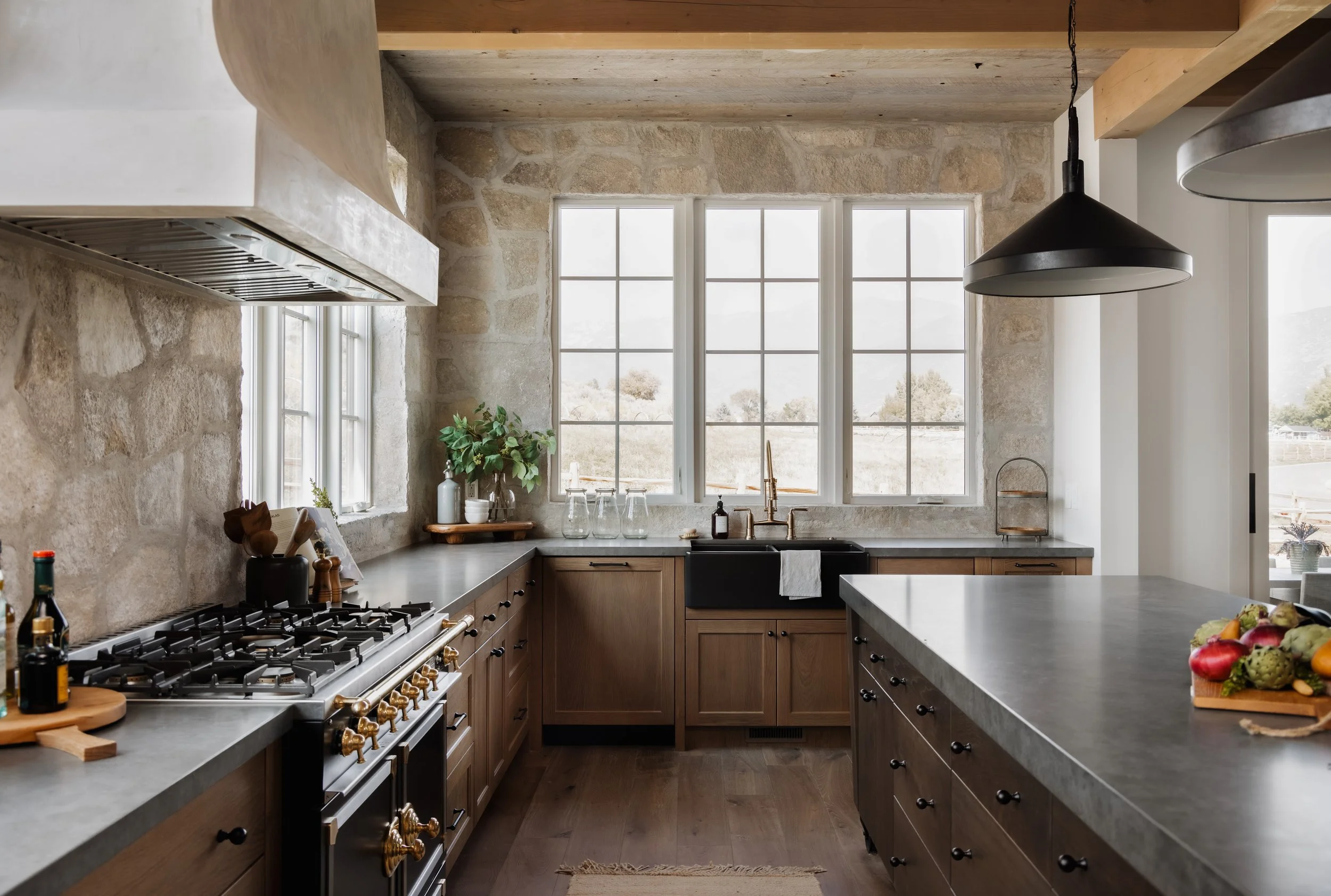 Kitchen with stone walls, large window, black farmhouse sink, wooden cabinets, and gray countertops. Includes a black stove with brass knobs, green plants, and fresh fruits on the island.