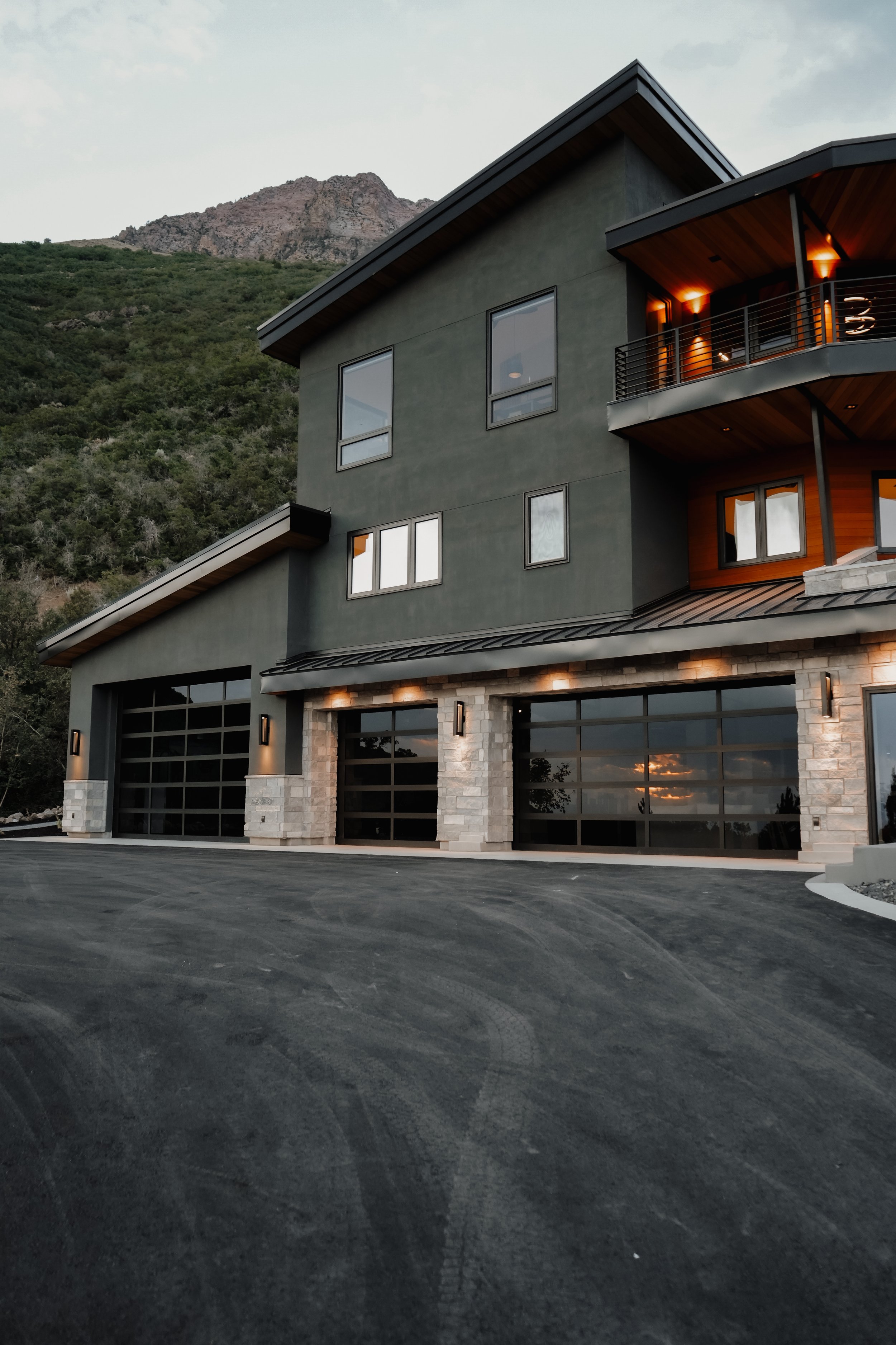 Modern multi-story house with green exterior walls and large windows, situated at the base of a mountain, with a paved driveway in the foreground.
