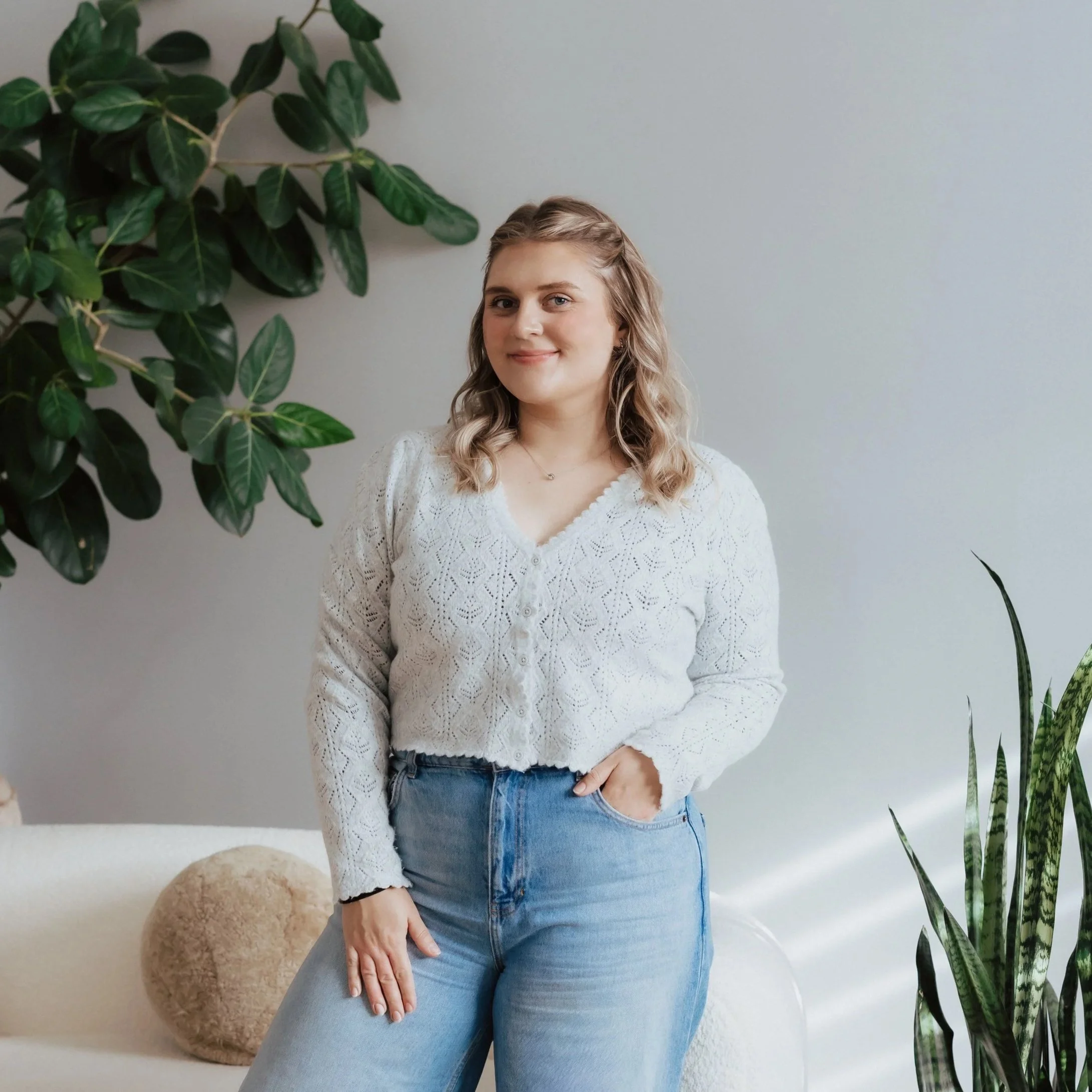 A young woman with curly, shoulder-length hair, wearing a white crochet sweater and light blue jeans, standing indoors in front of a plain wall and surrounded by green houseplants, smiling softly.