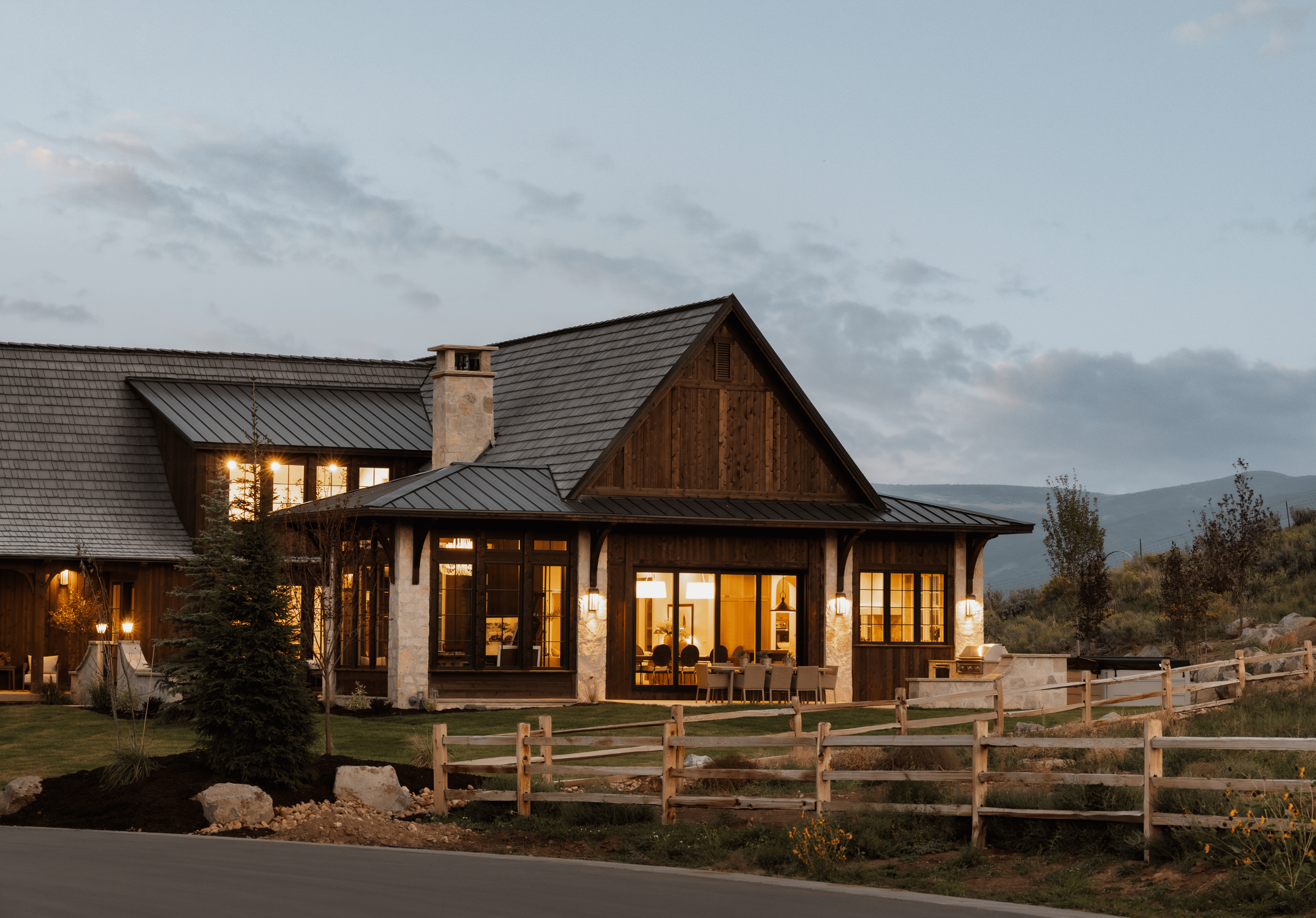 A house with a stone chimney, large windows, and a wooden exterior, illuminated from within, situated in a rural landscape with mountains in the background.