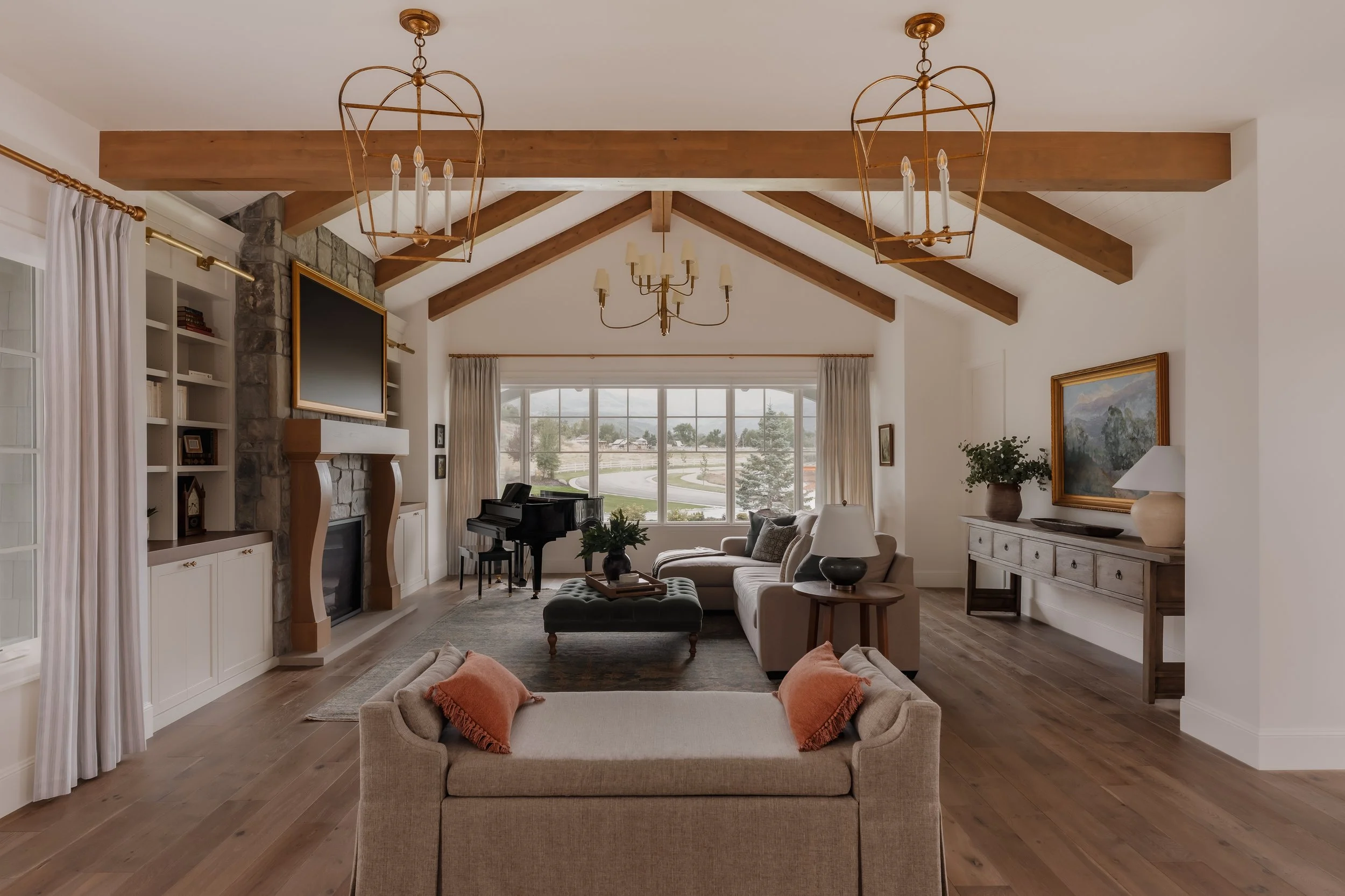 A spacious living room with a vaulted ceiling featuring exposed wooden beams, two chandeliers, and a large window with curtains. The room has a stone fireplace with a mounted flat-screen TV, a black piano near the window, a white sofa with throw pill