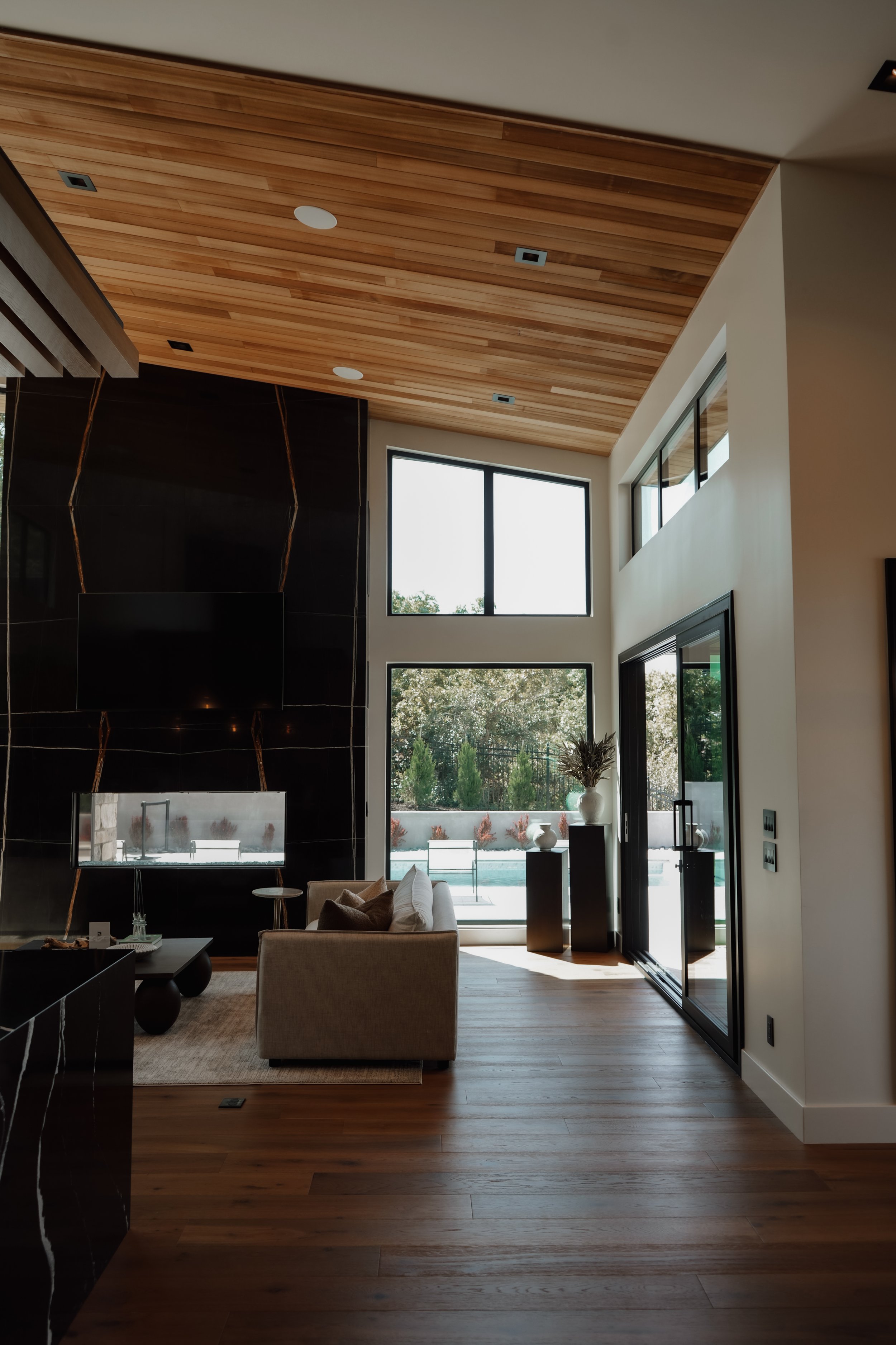 Modern living room with wooden ceiling and floors, black accent wall, large windows, and a beige sofa.