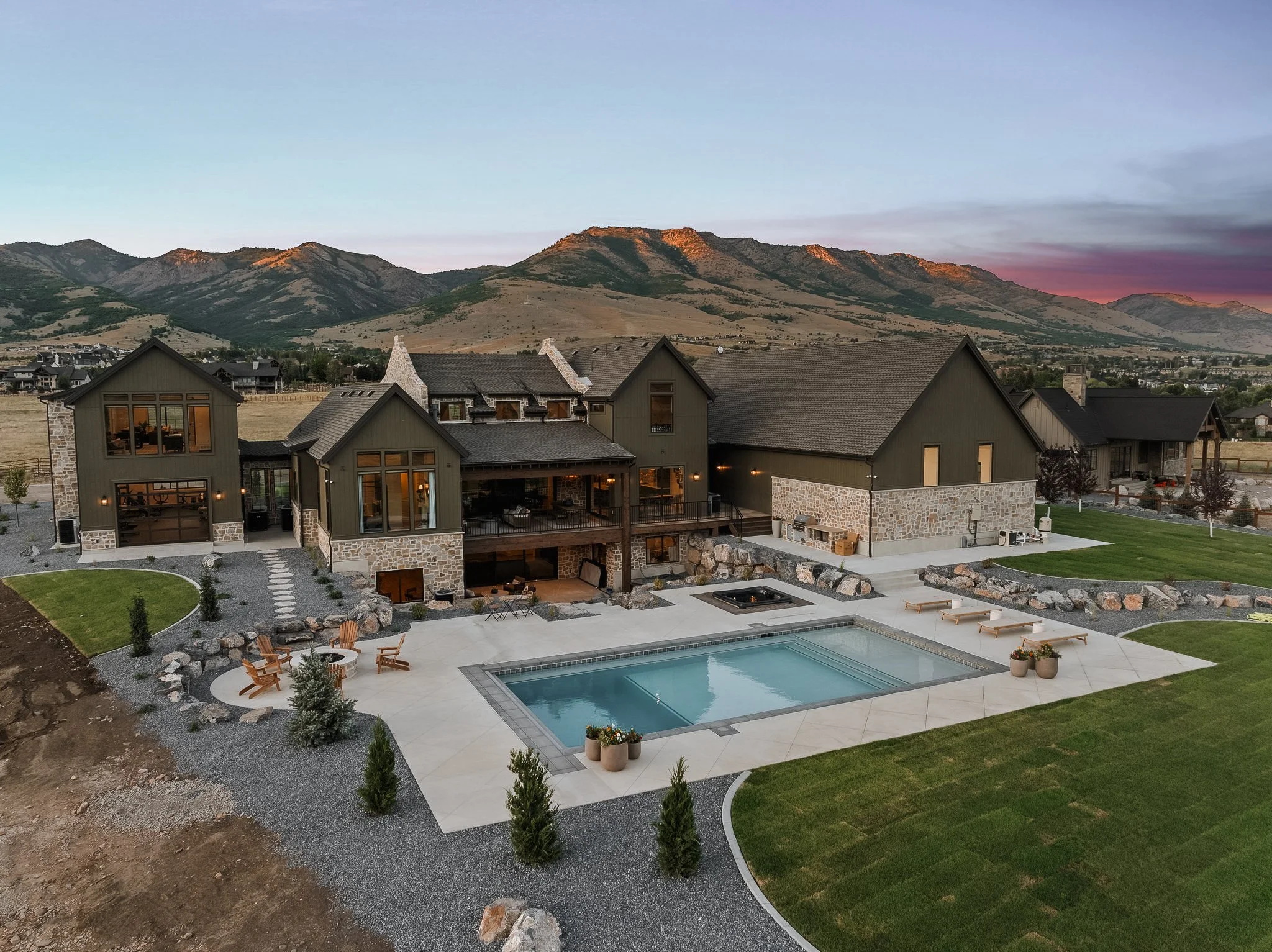 Large modern house with dark exterior and stone accents, landscaped yard, swimming pool with lounge chairs, fire pit, and mountain view in the background at sunset.