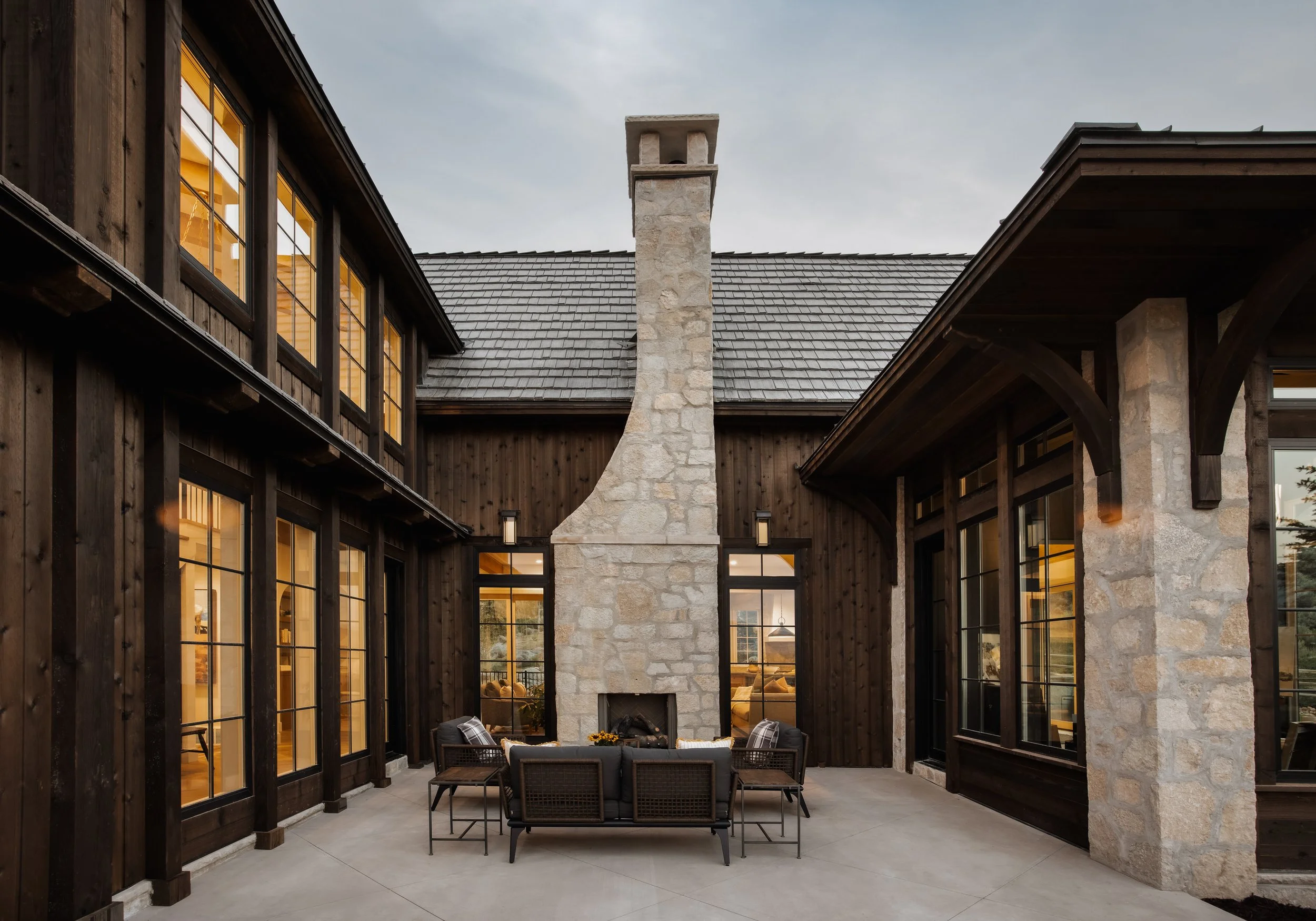 Outdoor patio area with a stone fireplace, dark wood siding, and large windows illuminated from inside, with outdoor seating and a concrete floor.