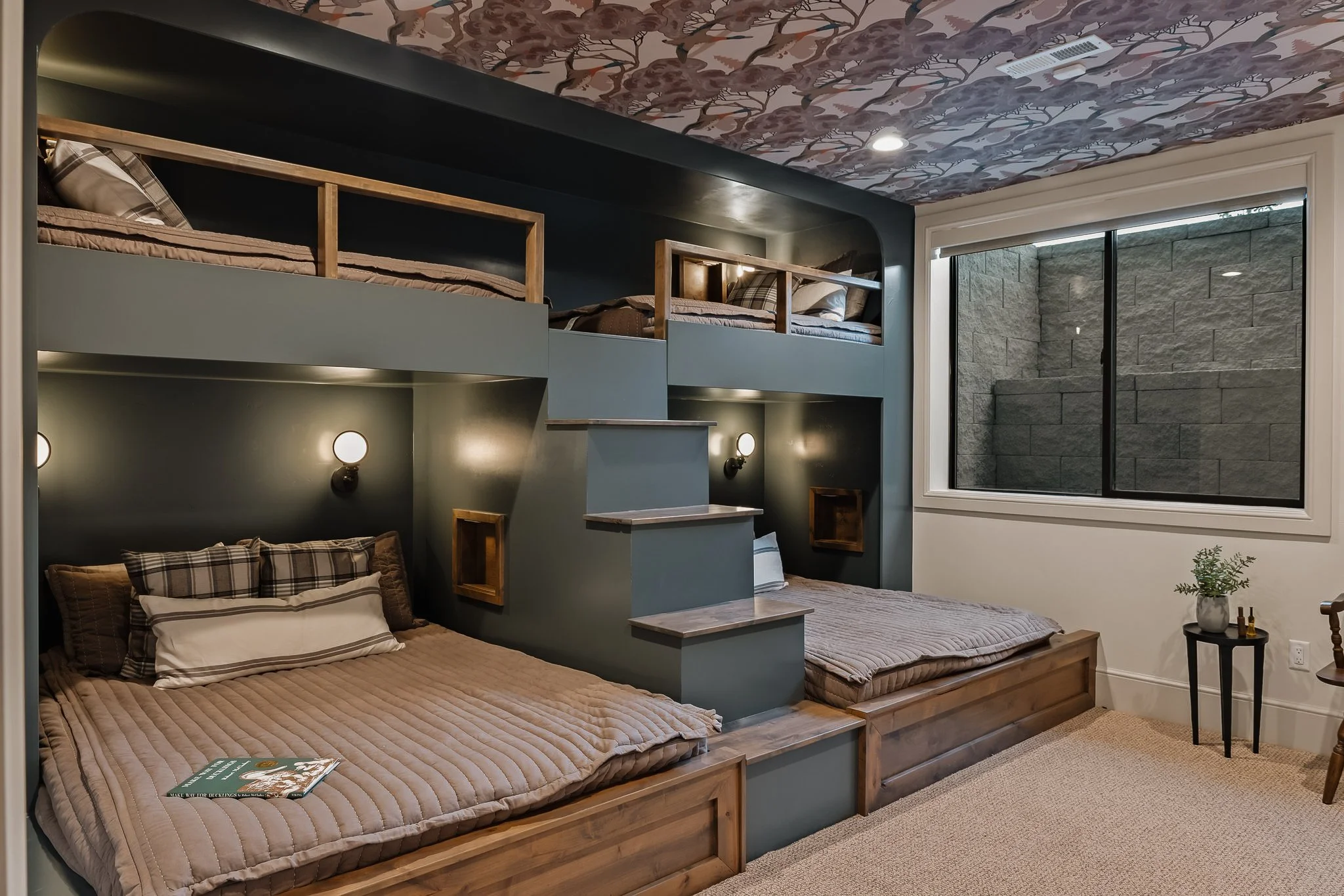 Room with two beds, one on the floor and one elevated. The room has dark green walls, wall-mounted lights, small open shelves, and a window with a stone wall outside.