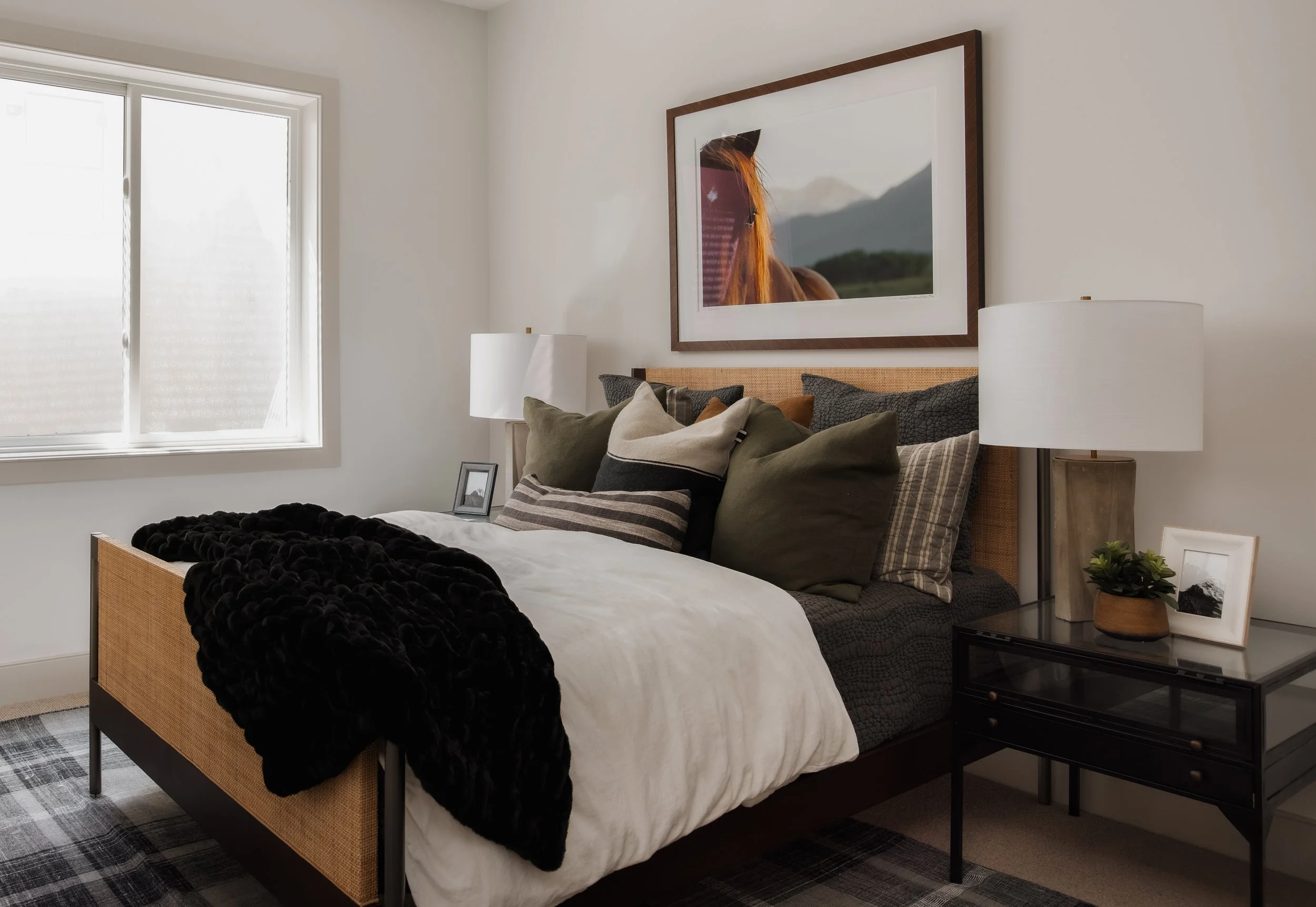 A cozy bedroom with a bed adorned with multiple pillows, a black throw blanket, and a framed horse photograph above the headboard. There are two white-lamped nightstands with framed photos and a small plant on the right side. A window with blinds let