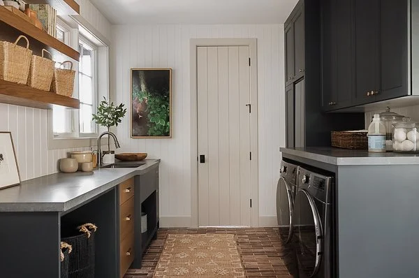 Laundry room with gray cabinets, a white door, a window with a plant view, a countertop with soap dispensers, laundry appliances, and open wooden shelves with baskets.