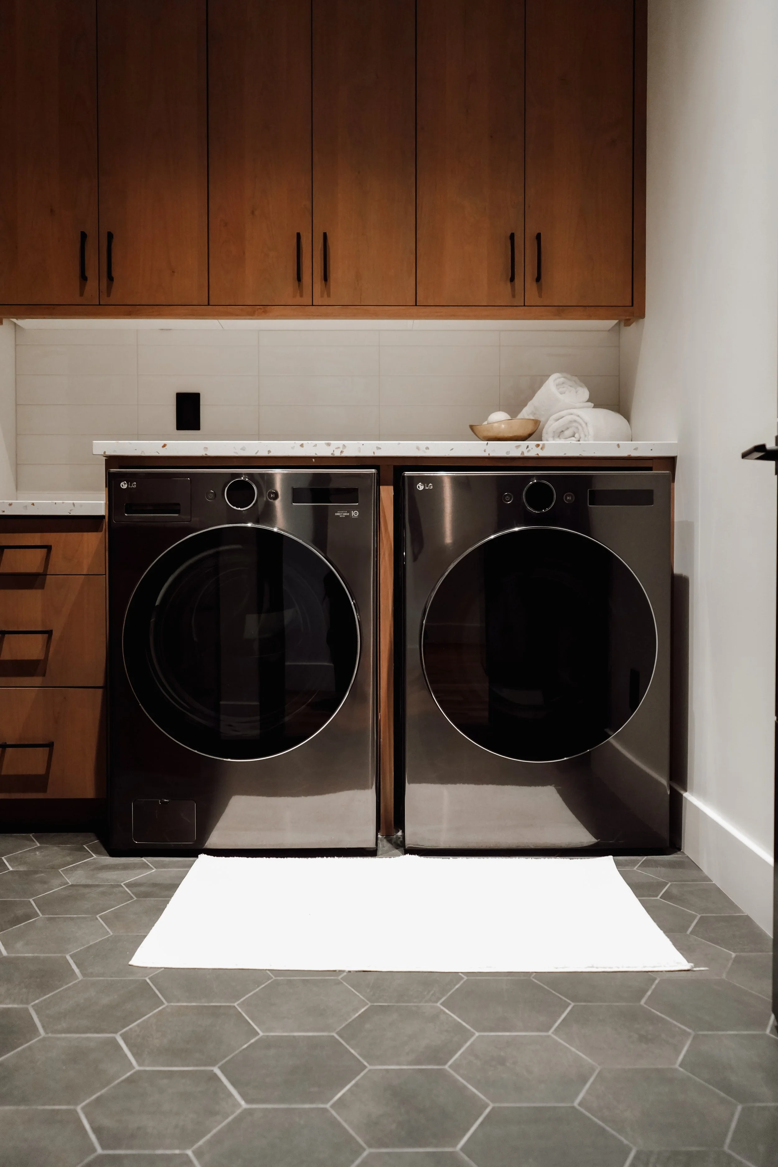 Stacked white towels on a small wooden bowl on top of a white counter between two black laundry washing machines in a modern laundry room with brown cabinets and gray hexagonal tiled floor.