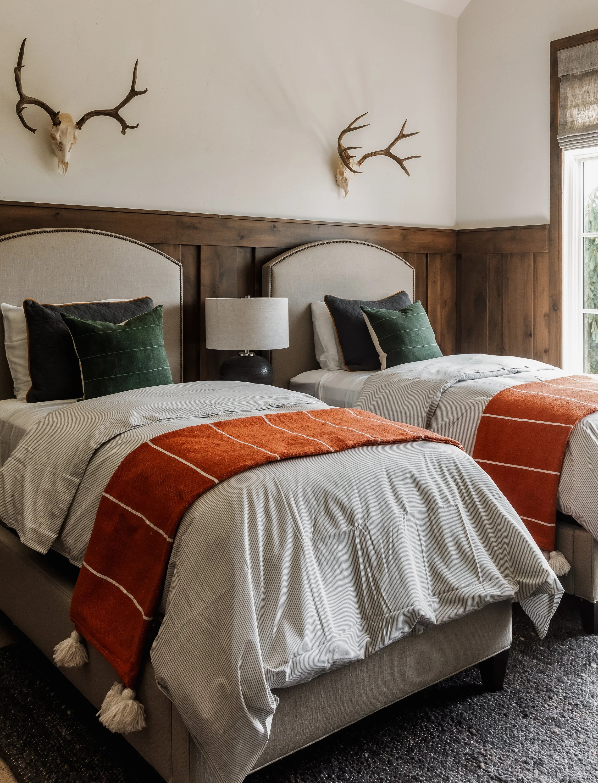 A bedroom with two beds, each with white bedding and orange and white striped throws. There is a nightstand with a table lamp between the beds, and deer skull with antlers mounted on the wall above each headboard. A window with blinds is on the right