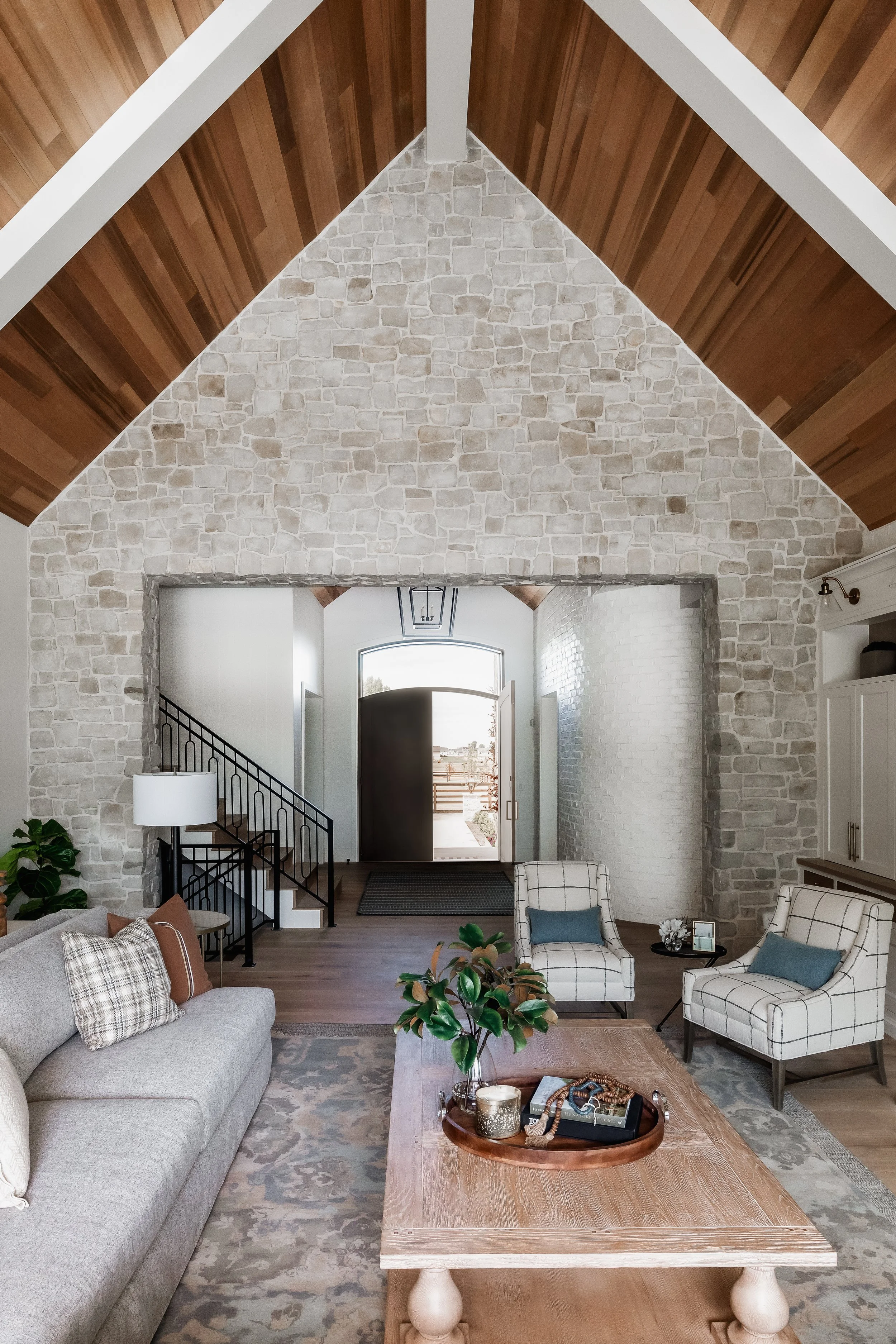 Living room with a stone archway, a wooden ceiling, a gray sofa with brown and plaid pillows, two white plaid armchairs with blue cushions, a wooden coffee table with a plant and decorative items, and an open front door leading outside.
