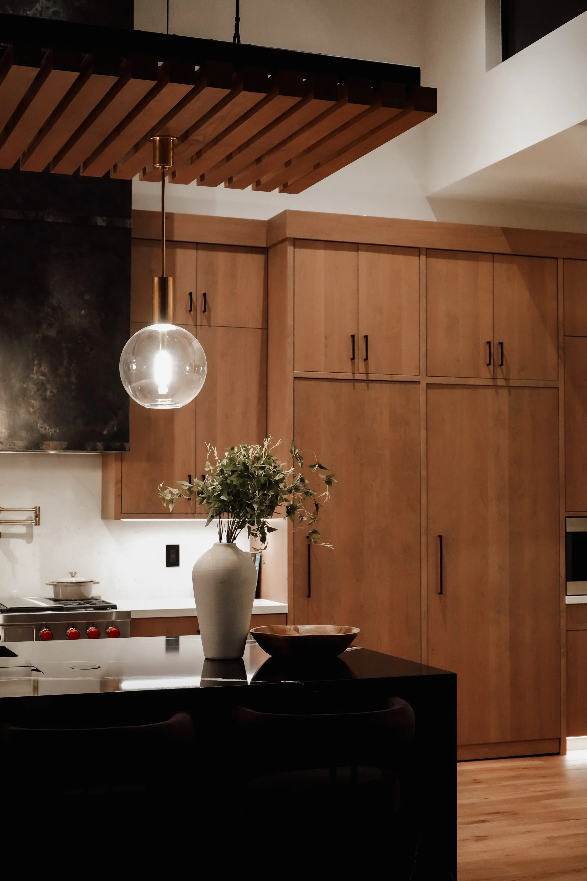 Modern kitchen with wooden cabinets, a black countertop island with a vase of greenery and a bowl, stainless steel stove, and a round glass pendant light.