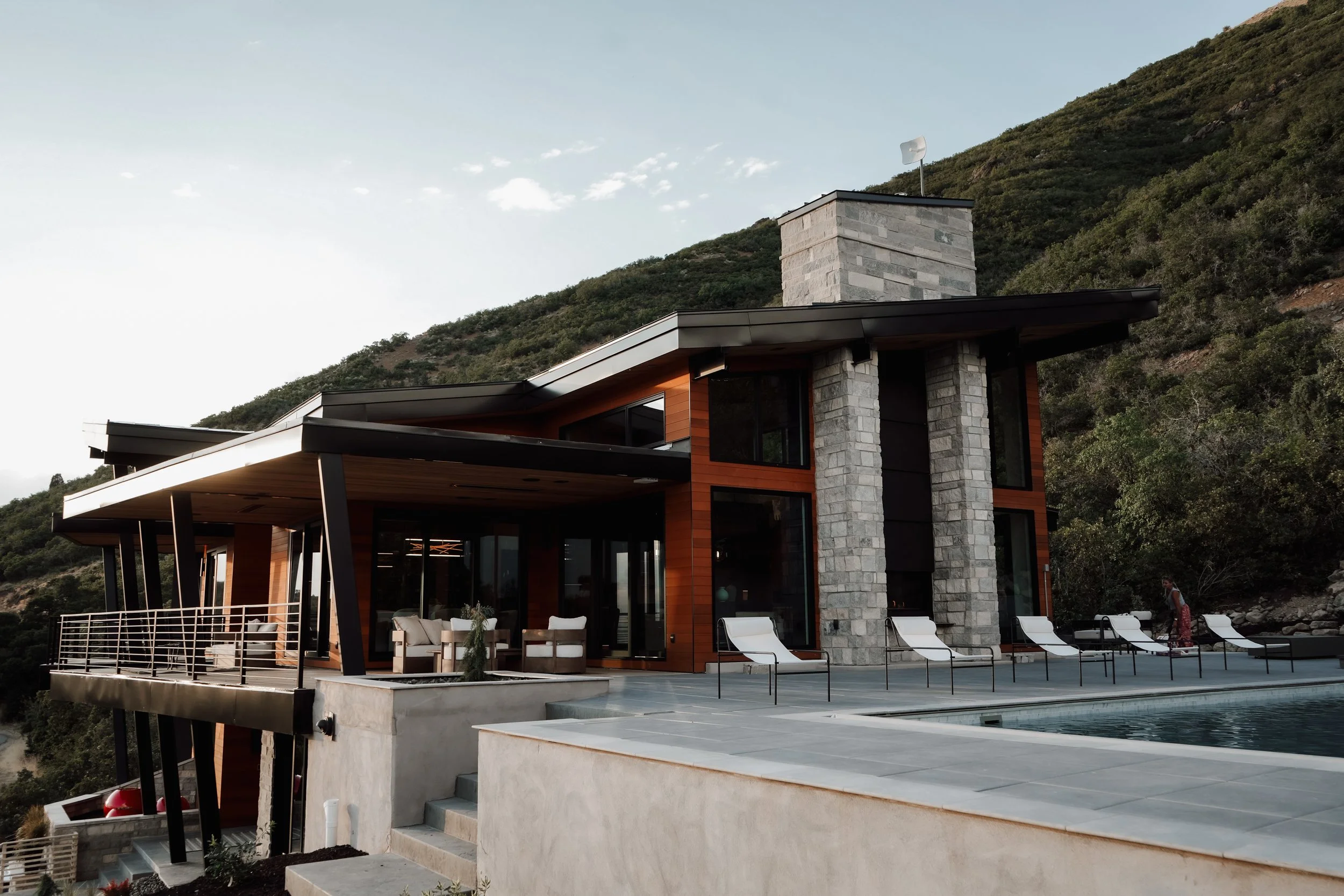 Modern house with large glass windows, stone chimney, and outdoor patio with lounge chairs, overlooking a hillside mountain.
