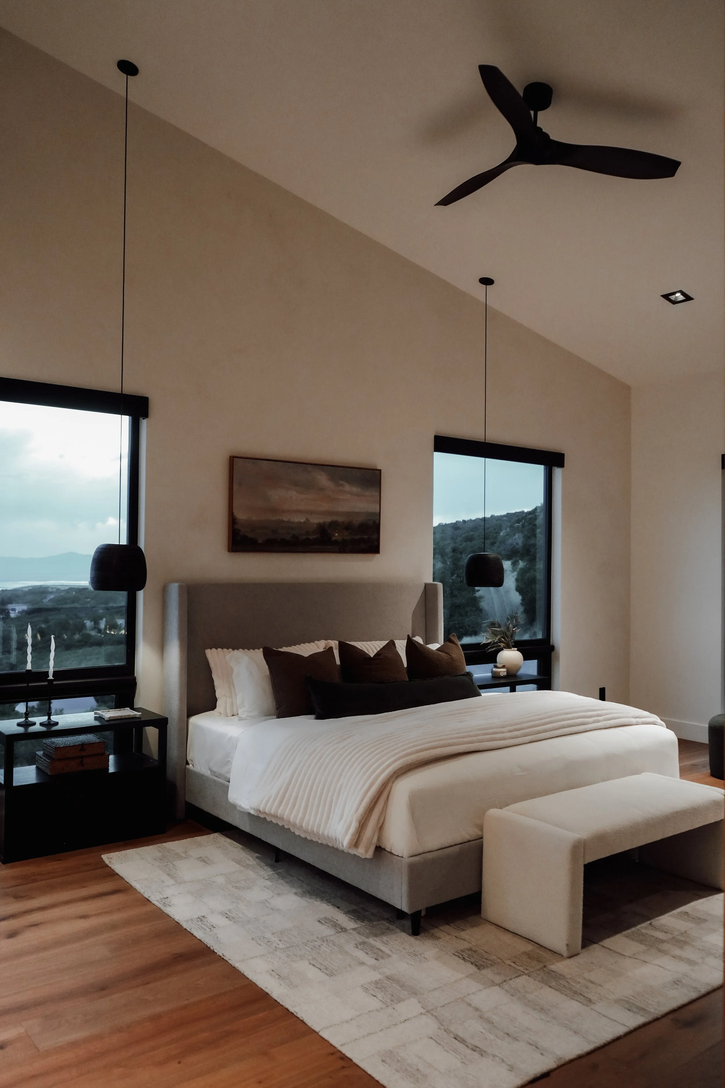 Modern bedroom with large gray upholstered bed surrounded by dark wood furniture, black pendant lights, and two windows overlooking a landscape of greenery and mountains.