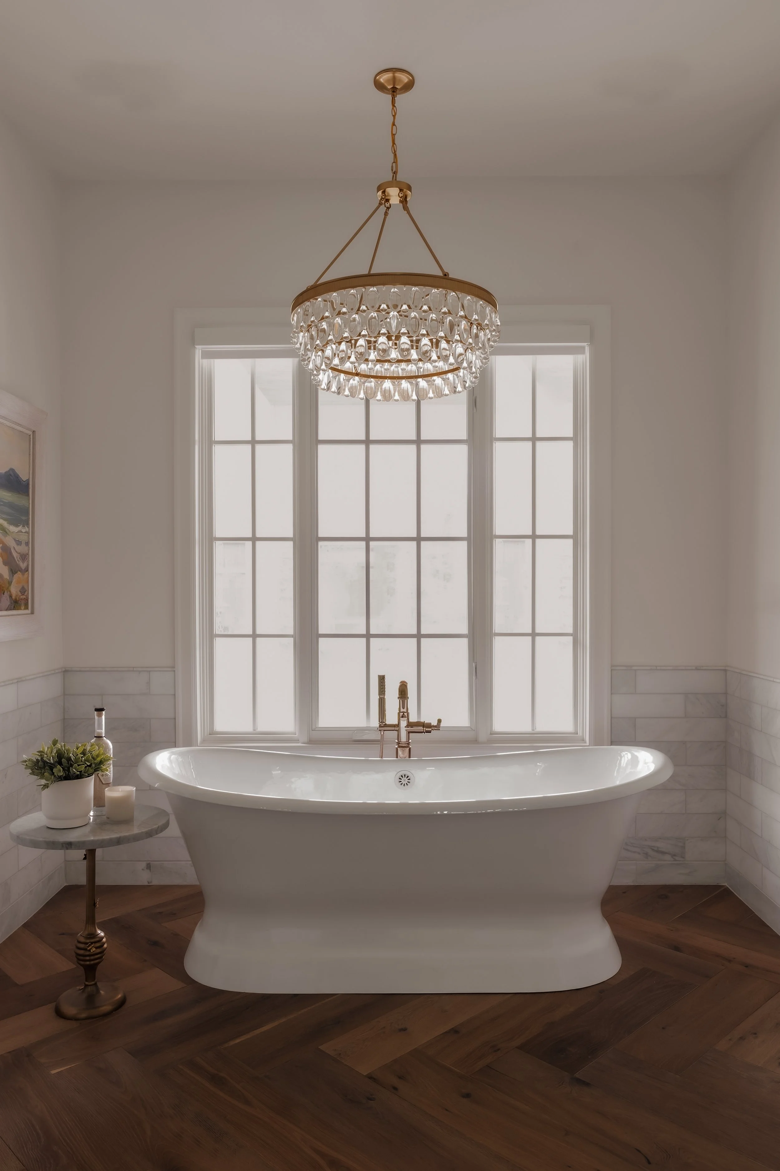 White clawfoot bathtub with a gold faucet, situated in front of large window with frosted glass, a small round side table with plants and candles, and a chandelier hanging from the ceiling.