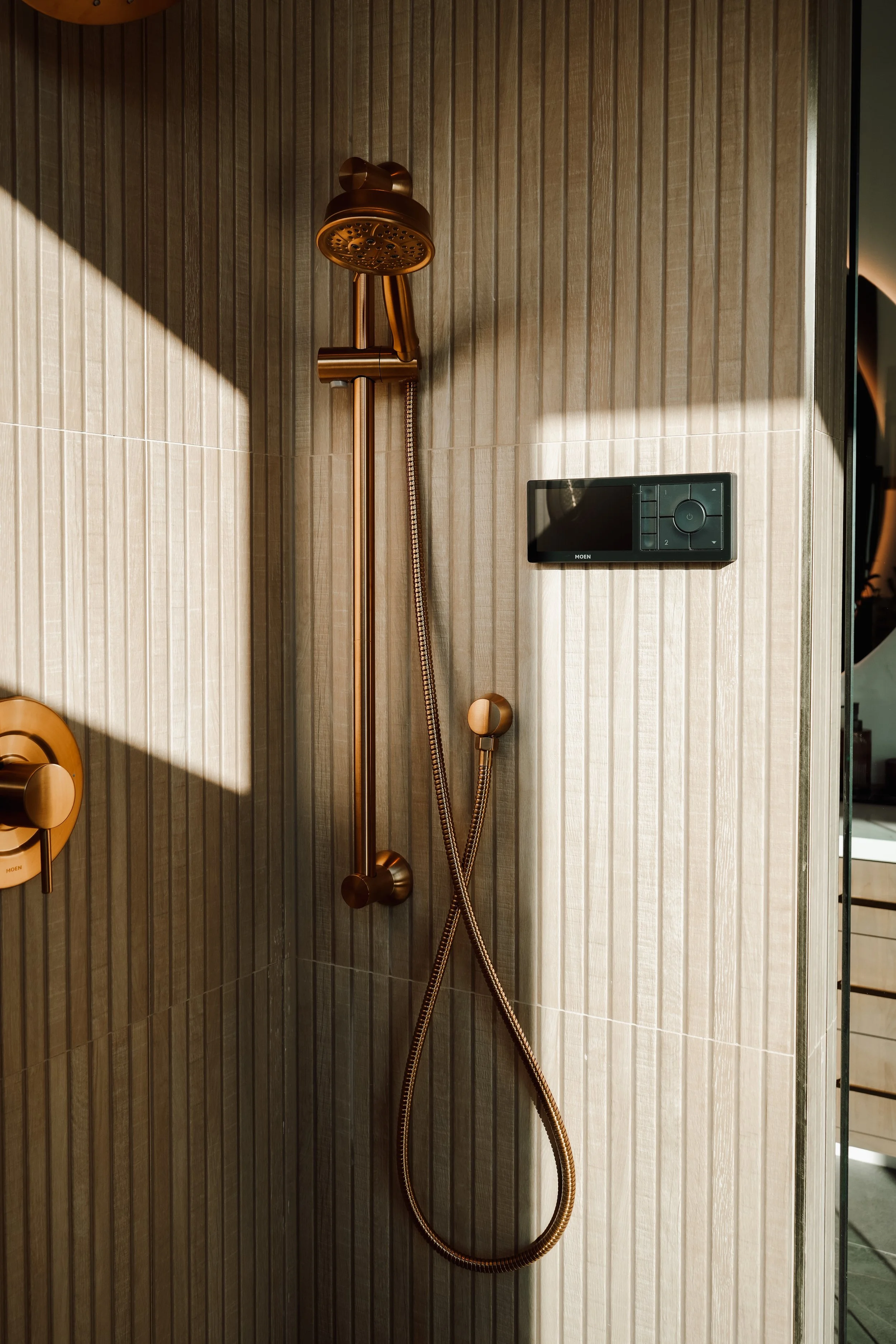 Modern shower with gold fixtures, a handheld showerhead, and a digital control panel on a beige tiled wall with vertical lines.
