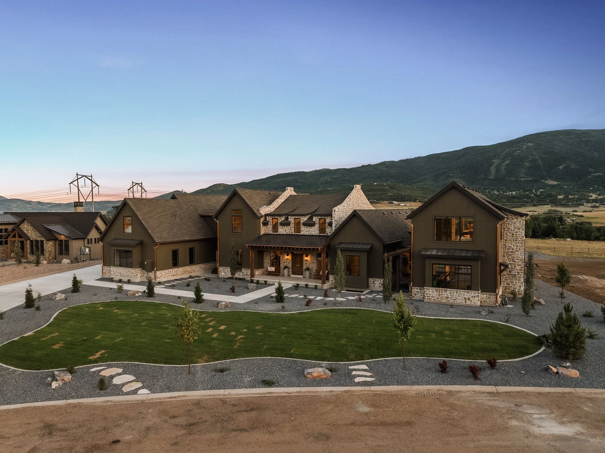 Exterior view of a modern residential house with landscaped front yard, paved walkways, small trees, and surrounded by scenic hills in the background during the evening.