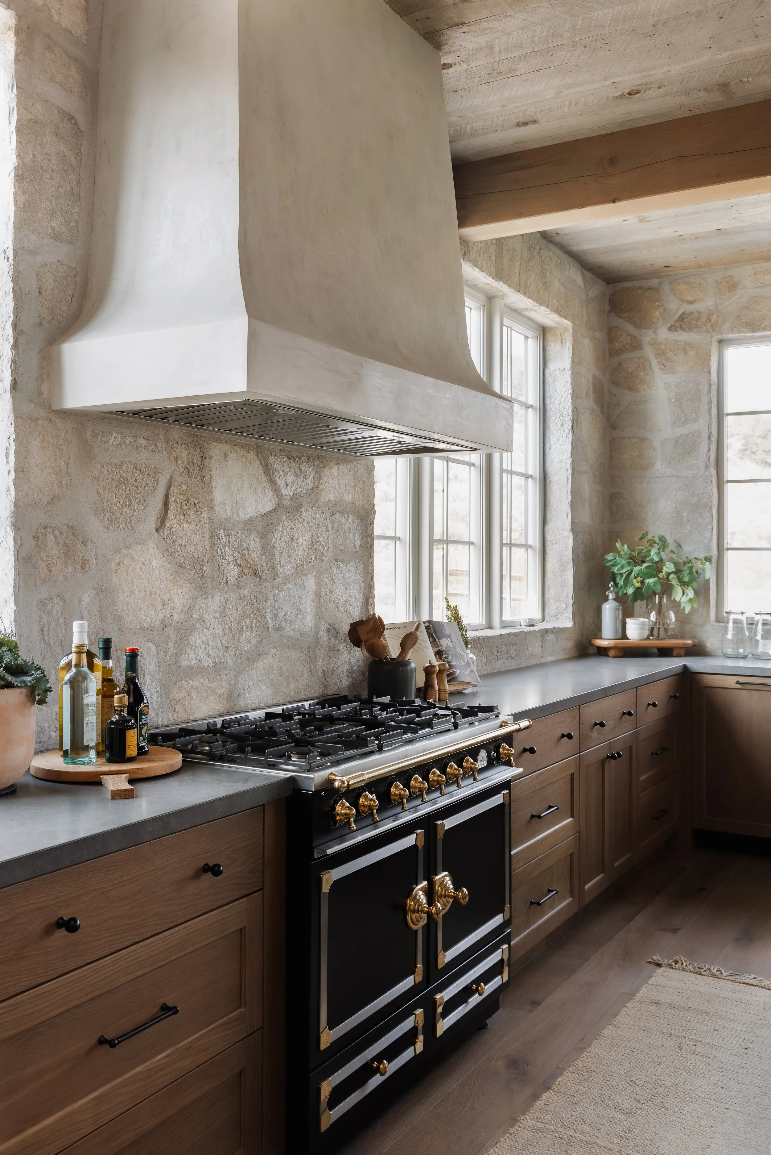 A rustic kitchen with stone walls, wooden cabinets, a black stove with gold accents, a large white range hood, and windows letting in natural light, decorated with potted plants and bottles.