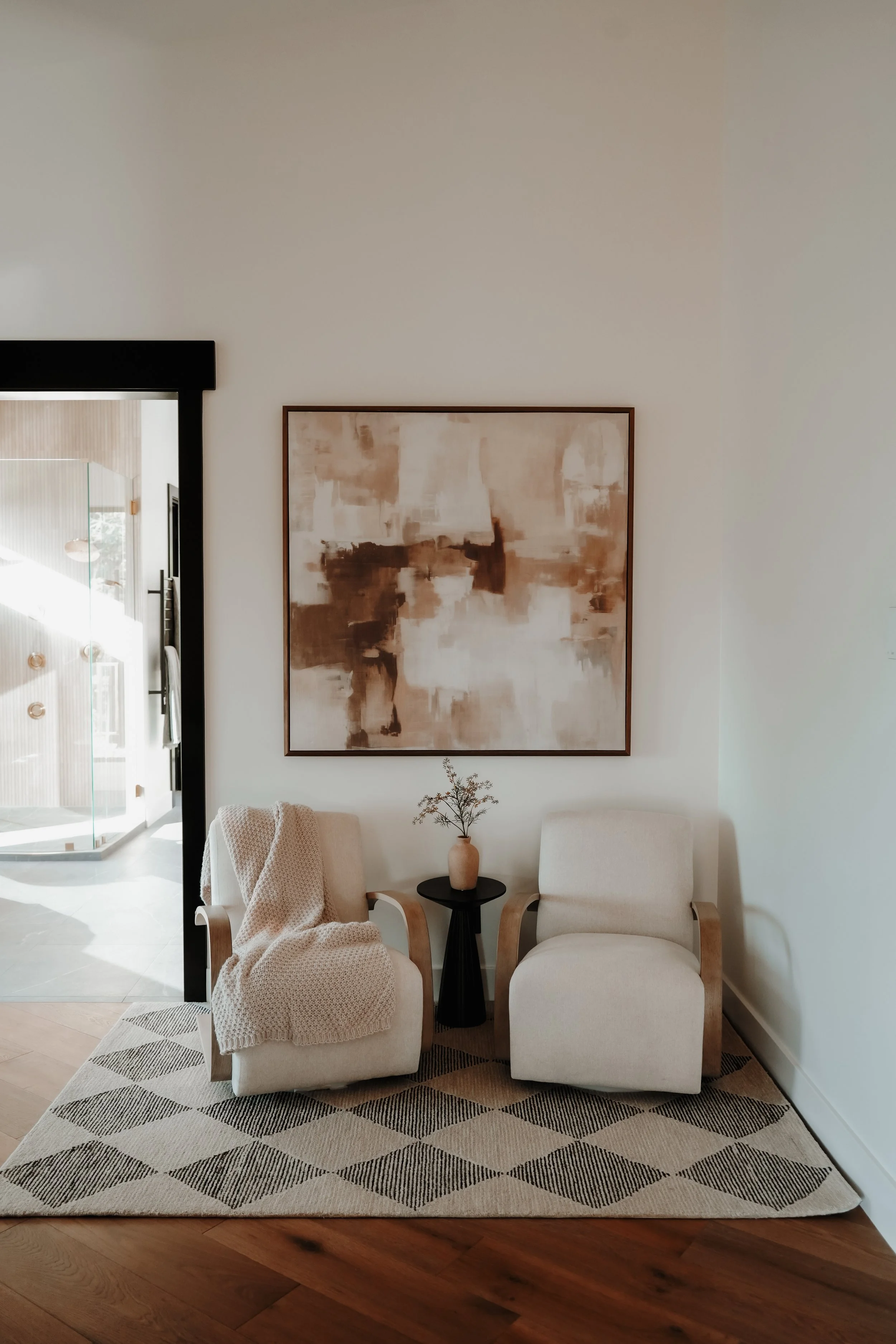 A cozy seating area with two white armchairs, a small black side table with a beige vase of flowers, a beige blanket draped over one chair, an abstract wall art piece, and a patterned rug on a hardwood floor.