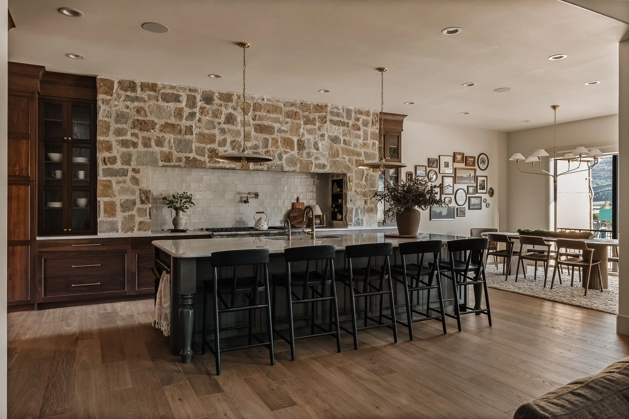 Open-concept kitchen and dining area with stone and wood features, black barstools at kitchen island, gallery wall of picture frames, large window with view, chandelier over dining table.