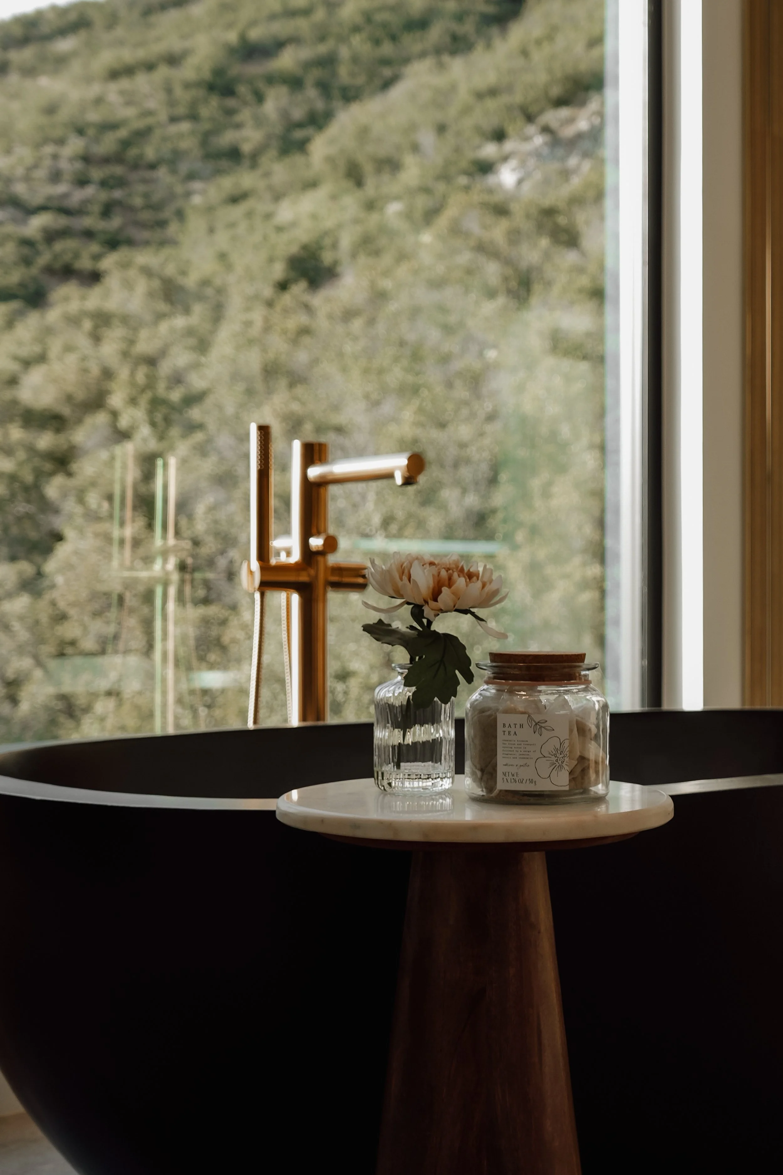 A black bathtub next to a window with a view of trees and hills outside. On a small round table near the tub, there is a vase with a flower and a jar labeled 'Bath Tea' with some bath salts inside.