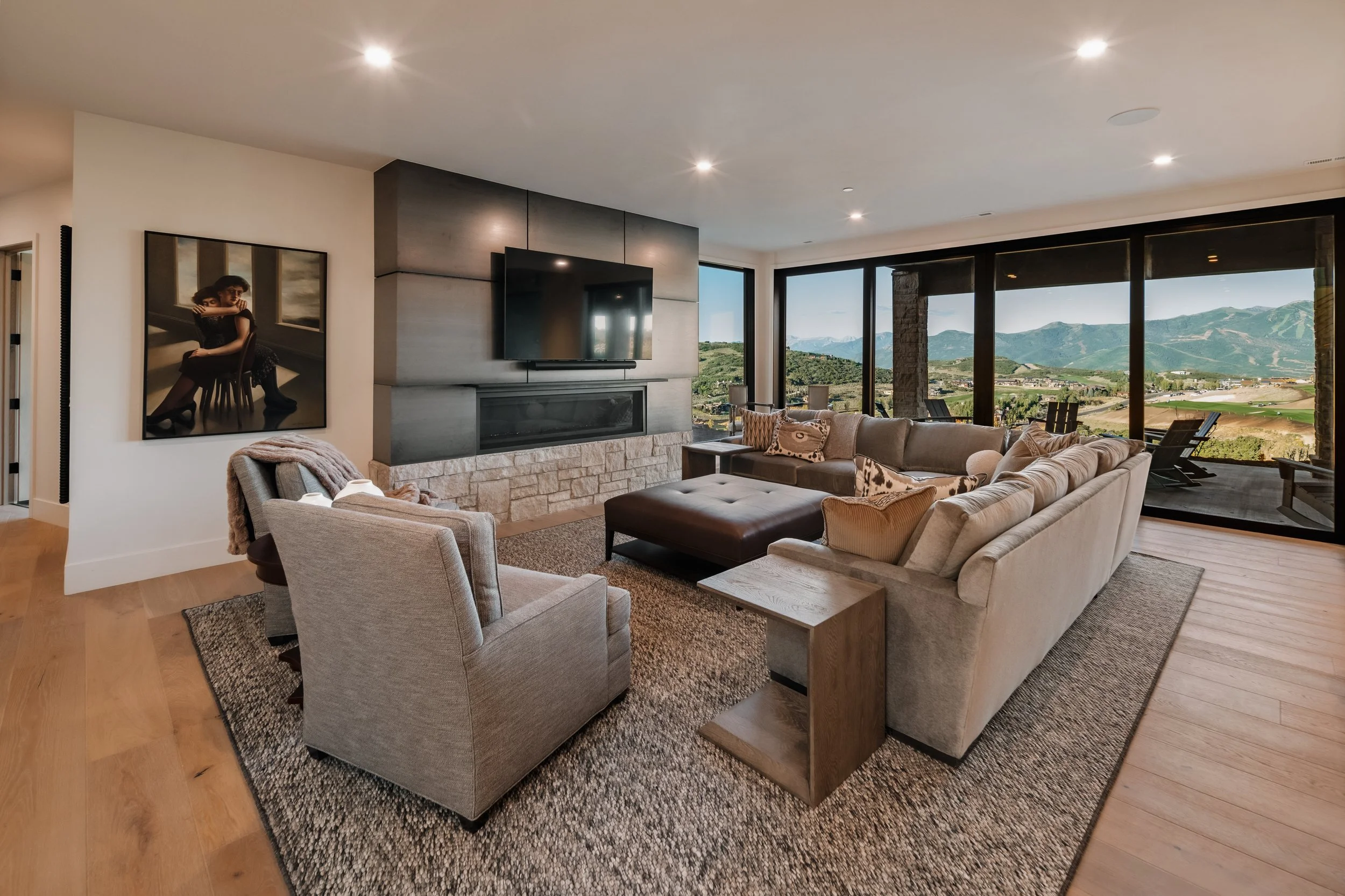 Modern living room with large beige sectional sofa, armchair, wooden side table, area rug, stone fireplace, wall-mounted TV, large windows showing mountain views, and outdoor seating area.