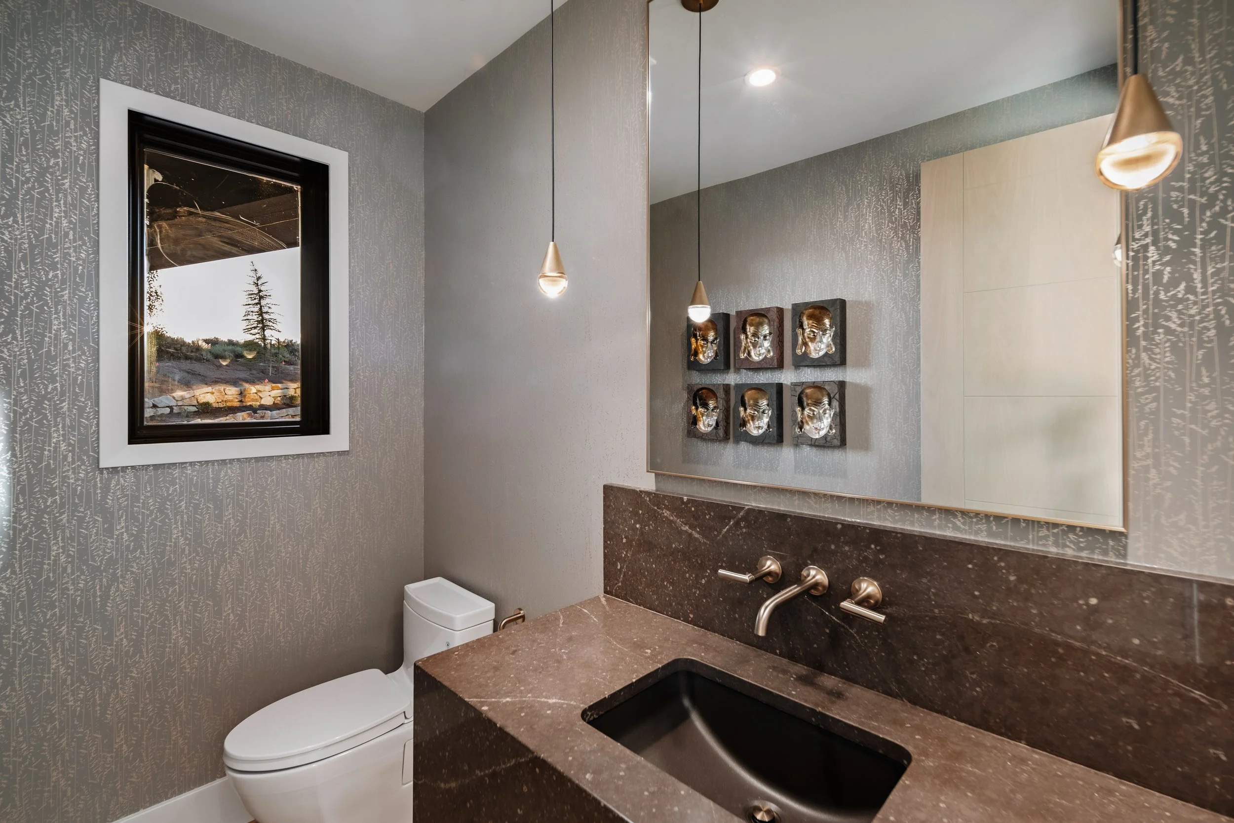 Modern bathroom with textured gray wallpaper, a small window, pendant lights, a wall art with metallic skulls, a dark brown marble countertop with a built-in black sink, a wall-mounted faucet, and a toilet.