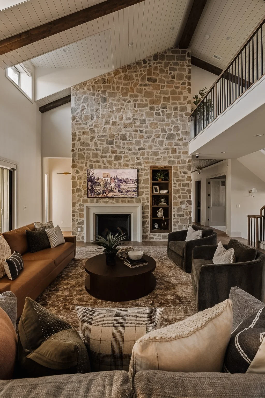 Living room with a high vaulted ceiling, stone fireplace, and a wall-mounted flat-screen TV. Contains sofas, armchairs, a round coffee table with a plant, and built-in shelves on the stone wall.