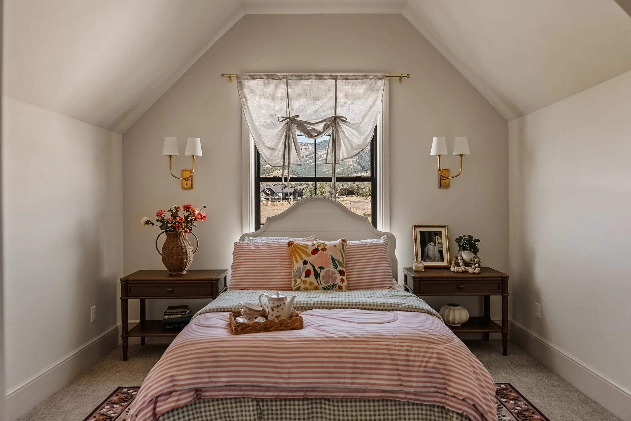 A cozy bedroom with a white framed bed, pink striped bedding, and a decorative pillow with floral design. The room has beige walls, a window with a striped curtain tied in the middle, two wall-mounted lamps, and matching wooden nightstands with decor