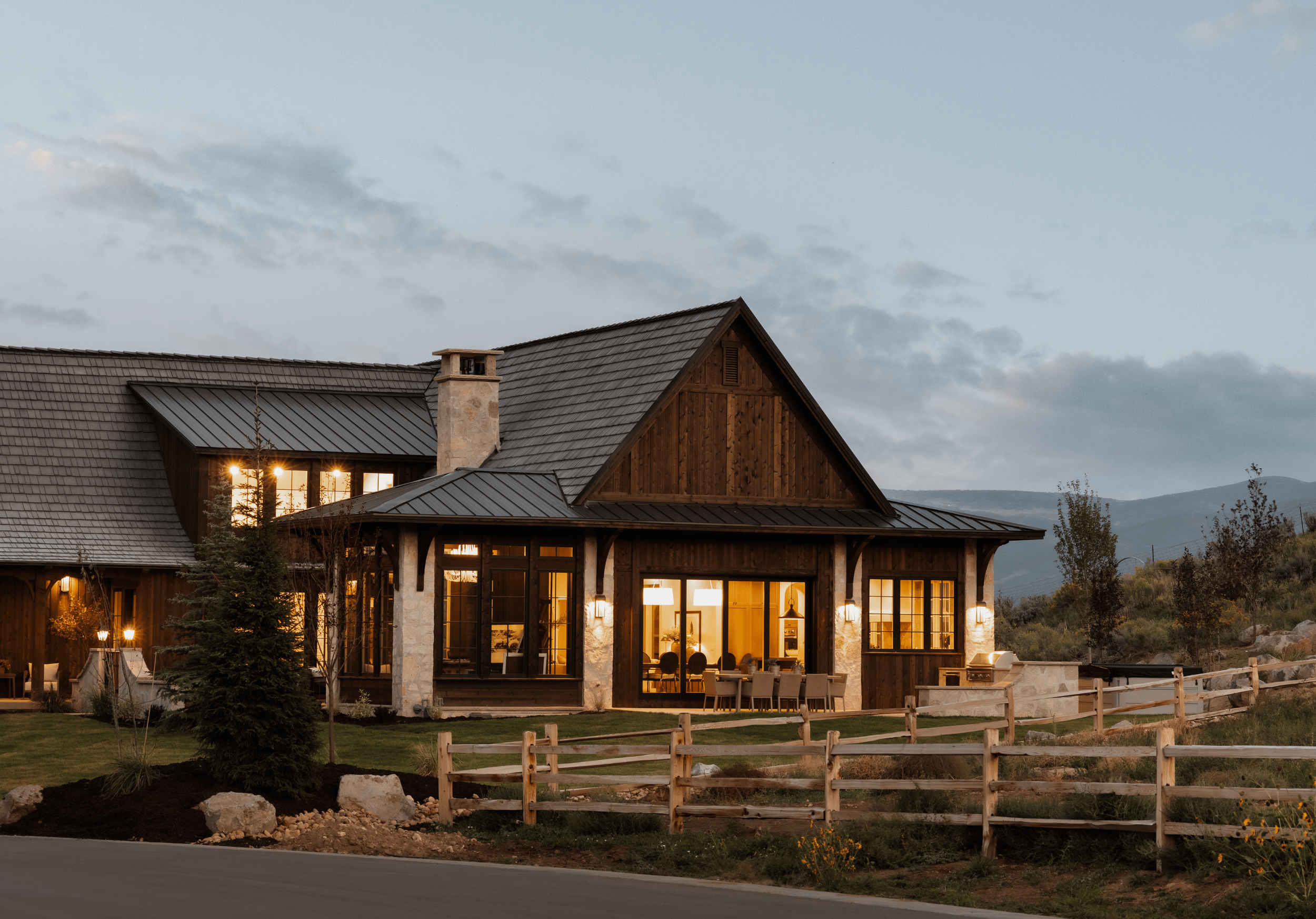 A large house with stone and wood exterior, lit from inside, surrounded by a wooden fence, with a mountainous landscape in the background at dusk.