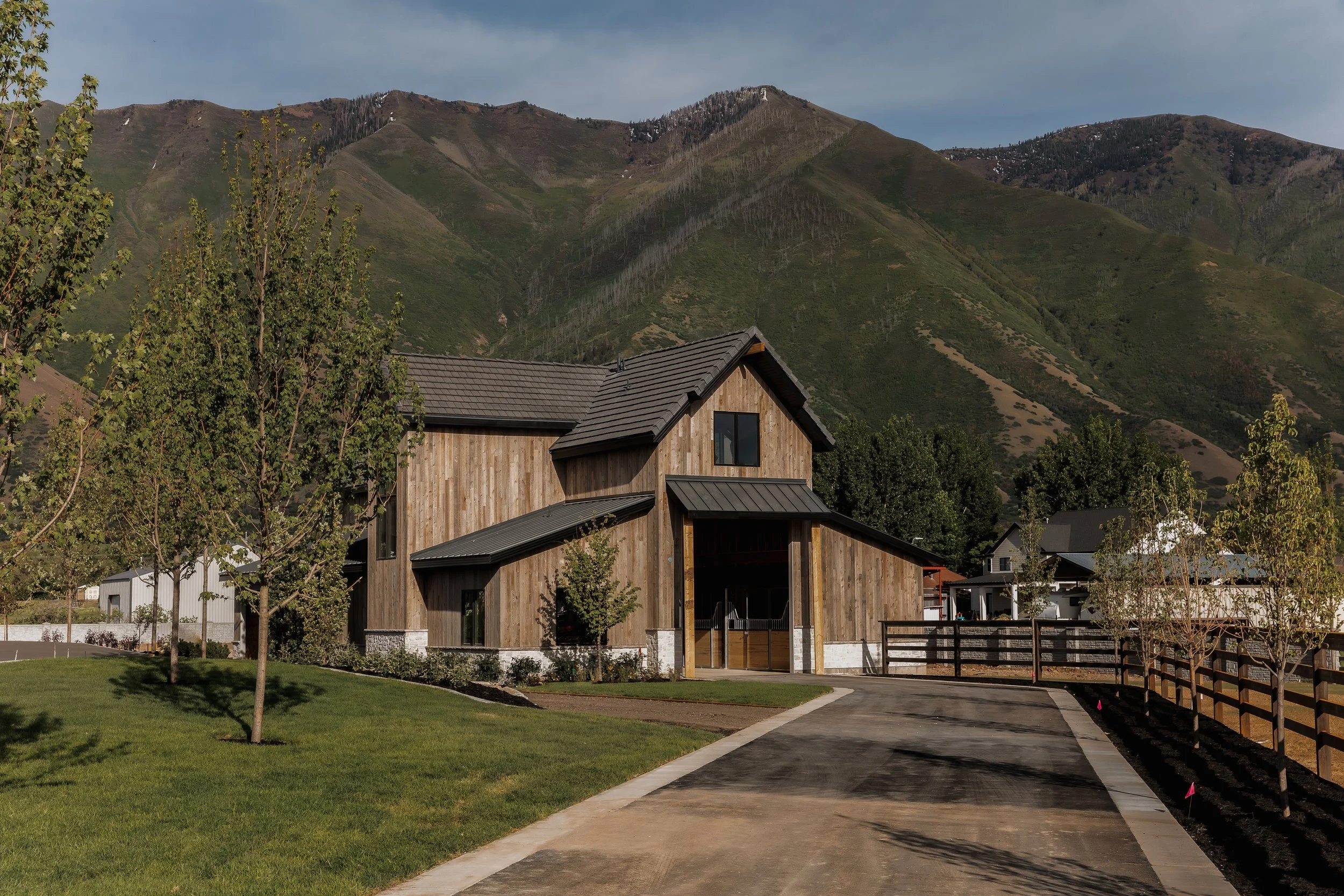A modern house with wooden siding and a dark gray metal roof, set against green mountains with patches of trees and snow. The house features a driveway and some small trees in the yard.