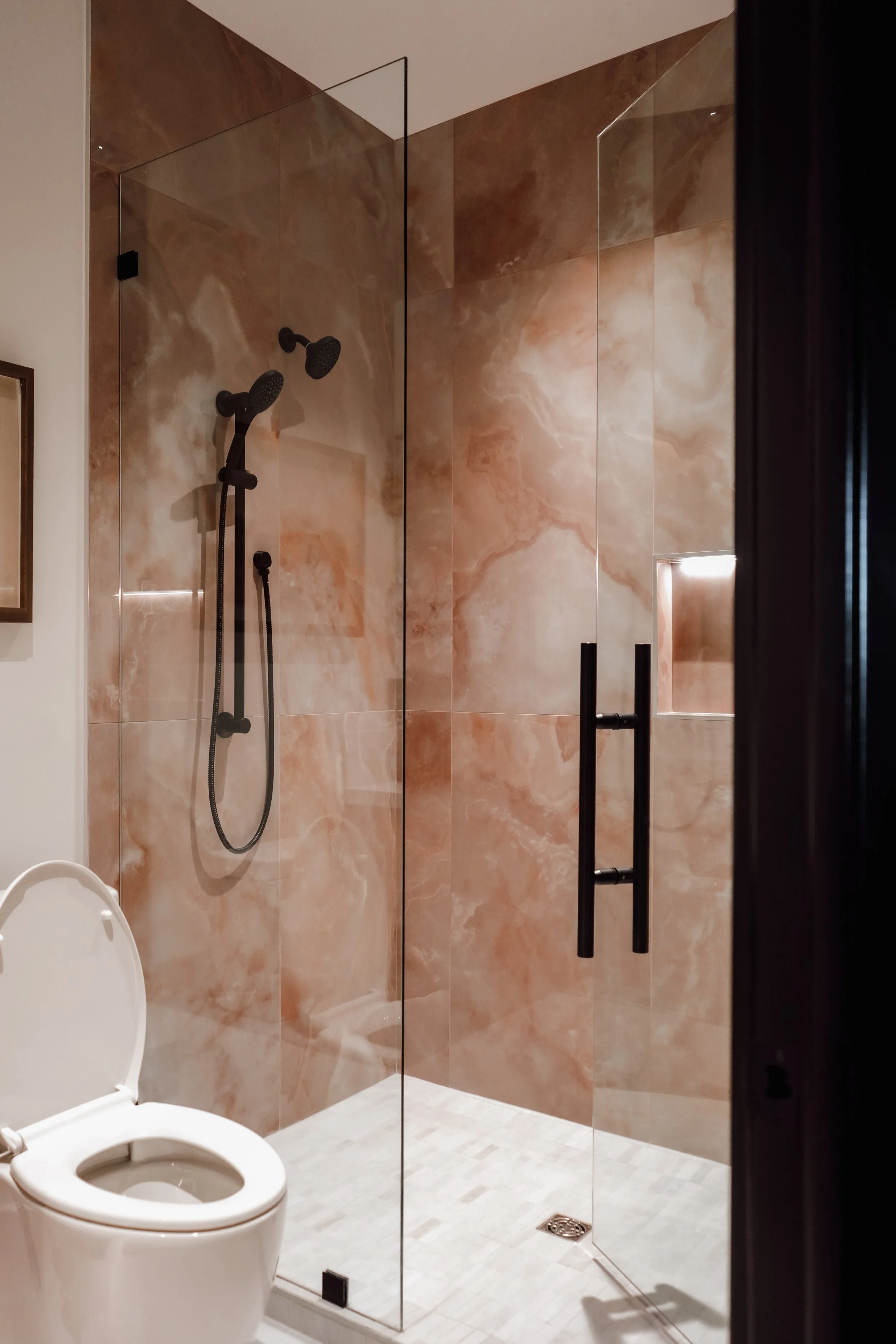 A modern bathroom with a walk-in shower enclosed by glass, featuring black fixtures including two showerheads and a handheld shower. The shower walls are covered in brown marble tiles, and there is a black towel bar on the door. A white toilet with a