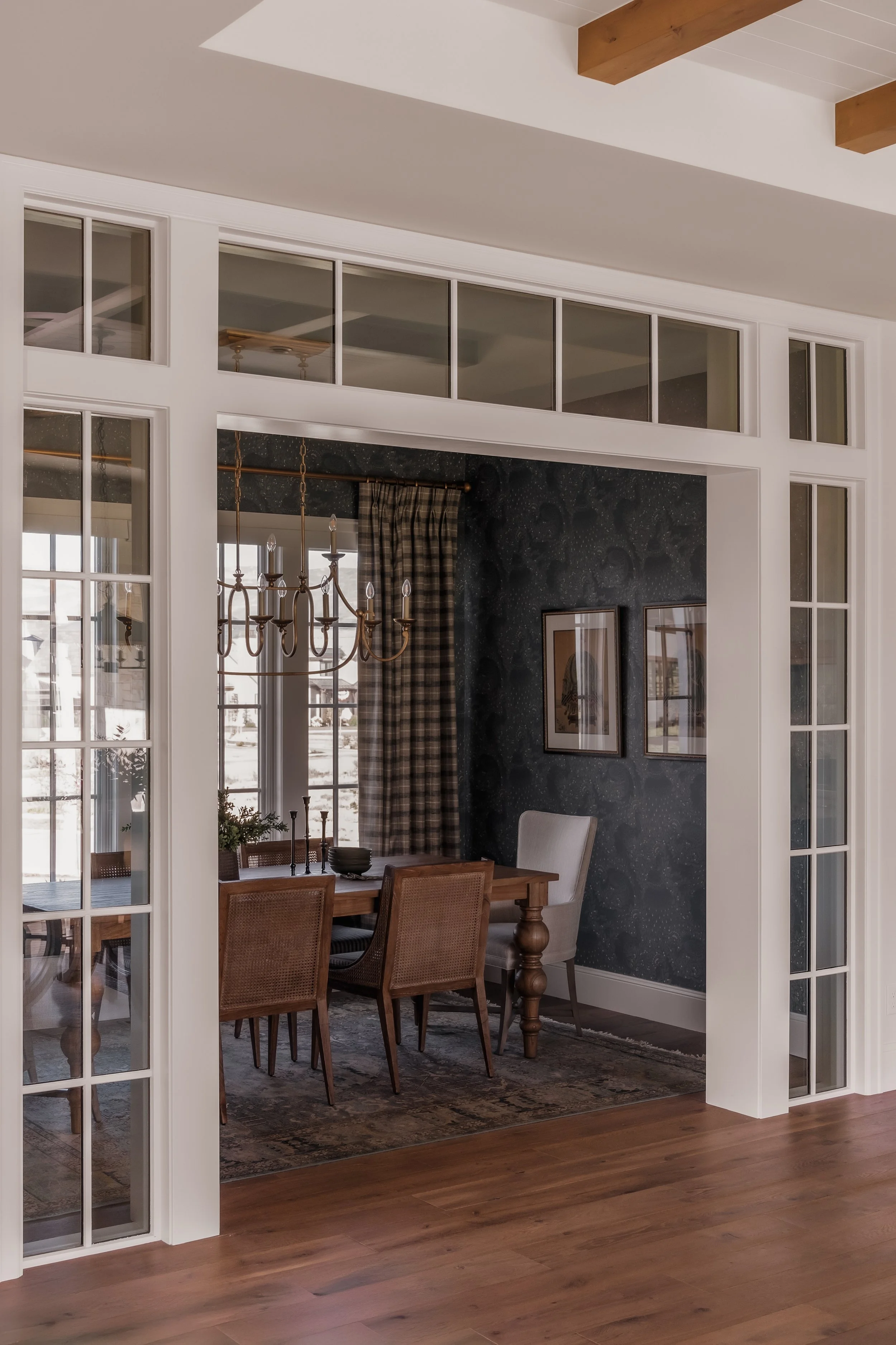View of a dining room seen through a large white-framed glass door and window, with a wooden table, chairs, a chandelier, and a dark wall with framed pictures.