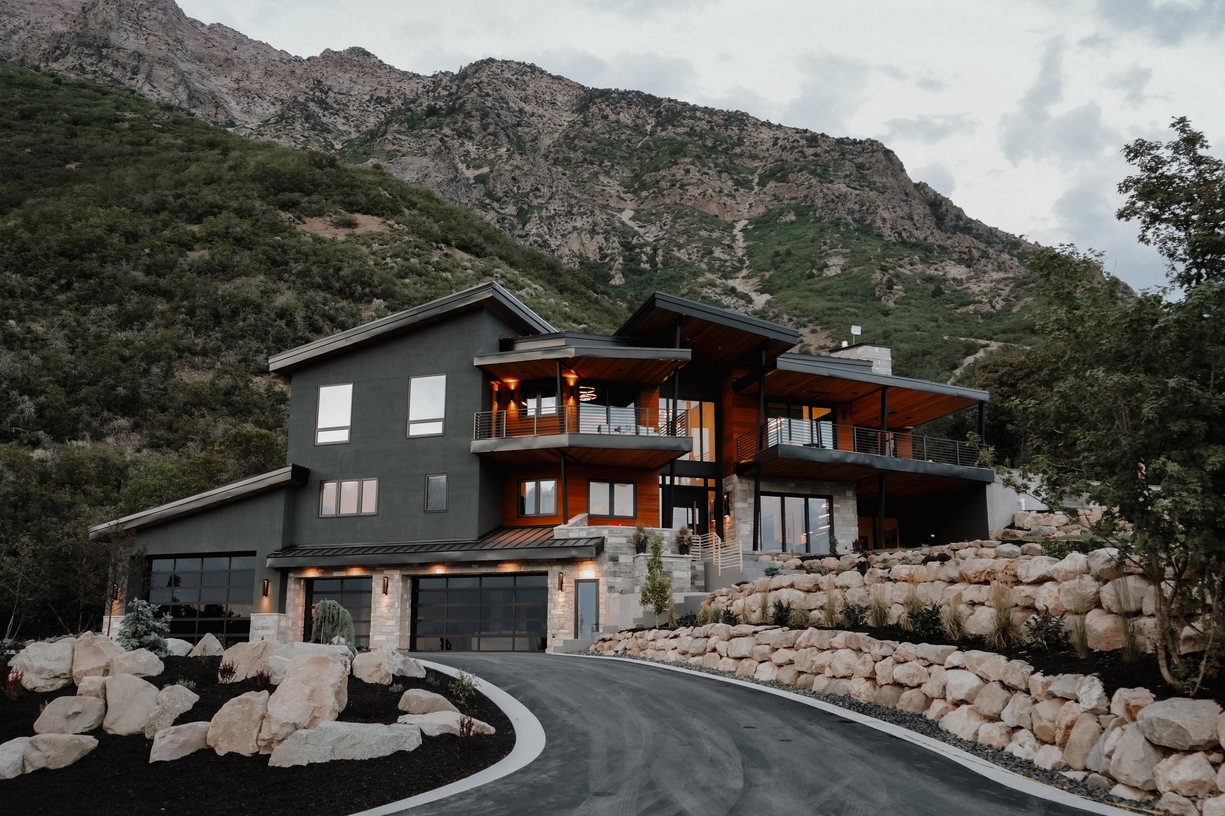 A modern multi-story house with large windows and balconies, situated on a hillside with a curved driveway and rocky landscaping, with mountains in the background.