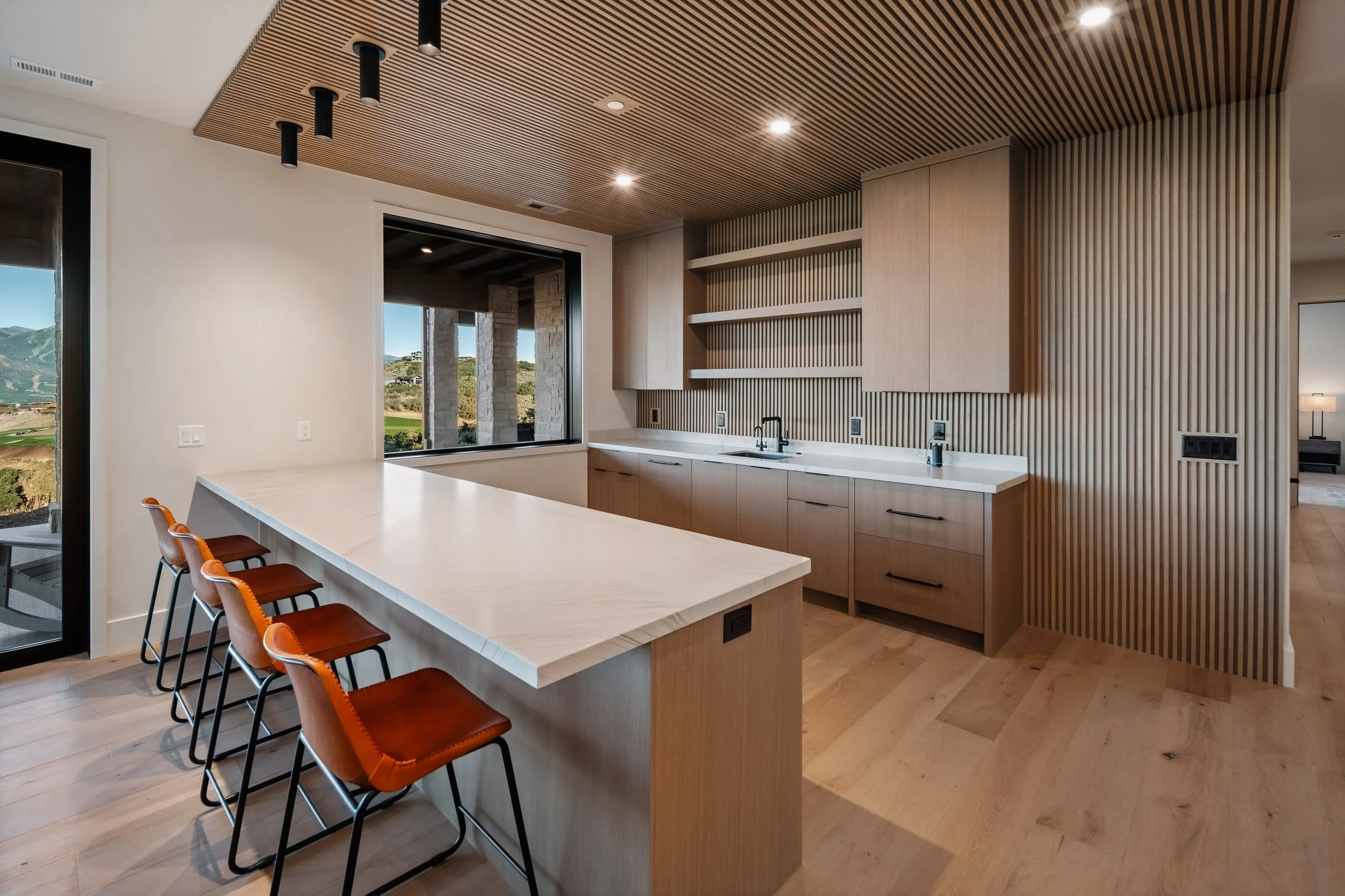 Modern kitchen with light wood cabinets, a white marble island with orange barstools, wood slatted ceiling, and large window showing outdoor landscape of mountains and greenery.