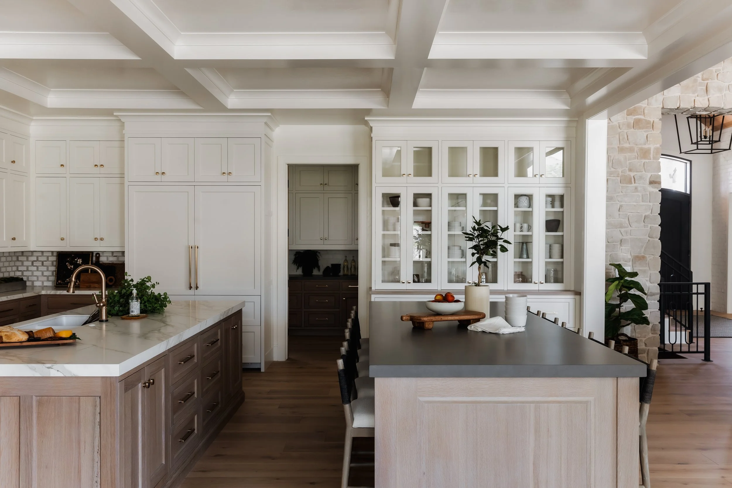 Modern kitchen with white and wood cabinetry, a large island with a dark gray countertop, and a built-in glass-front cabinet with dishes and decor. There are plants, bowls, and apples on the island, and a stone accent wall on the right.