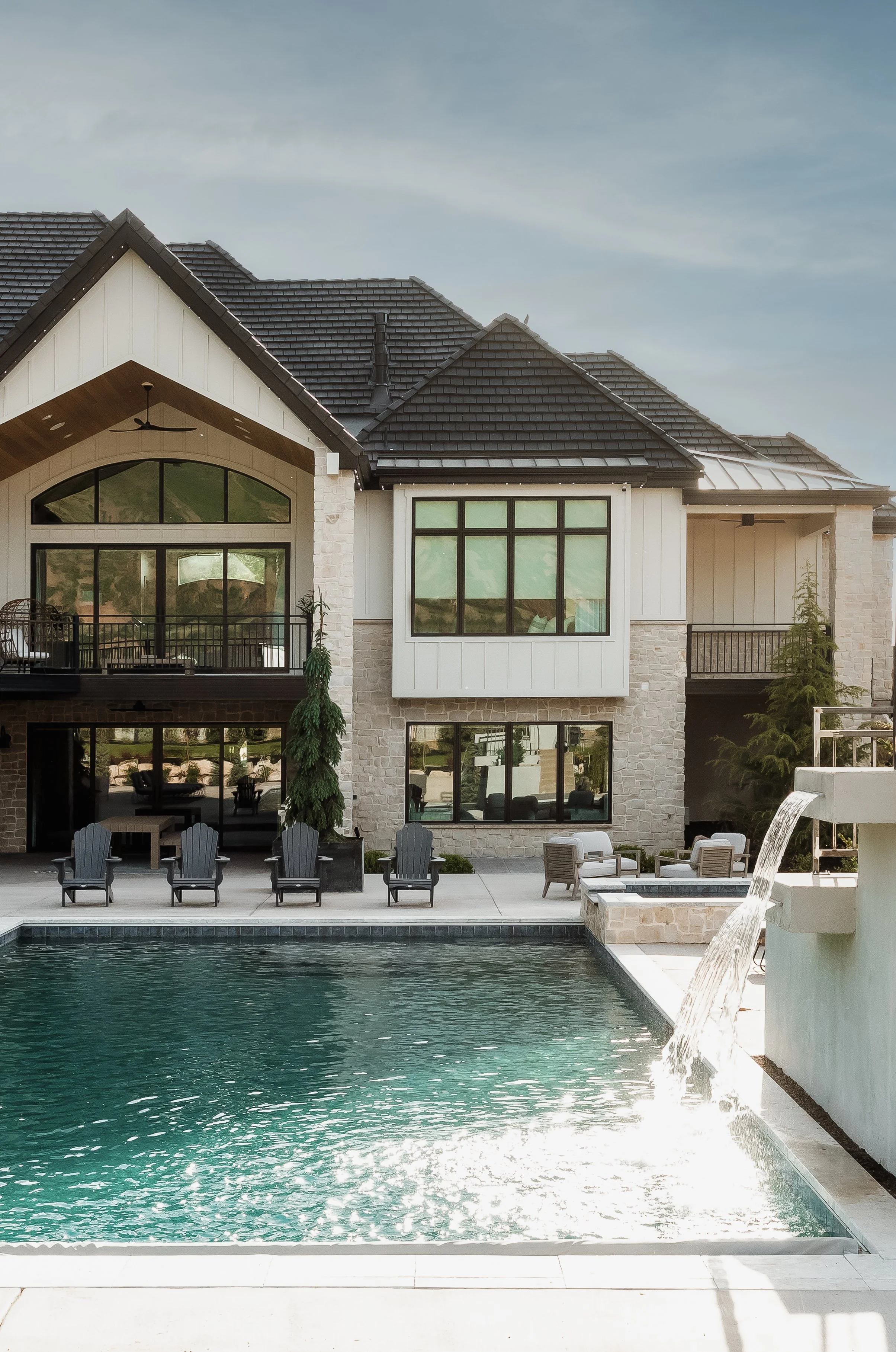 A modern house with a swimming pool in the foreground, outdoor chairs, a waterfall feature, and lush greenery.