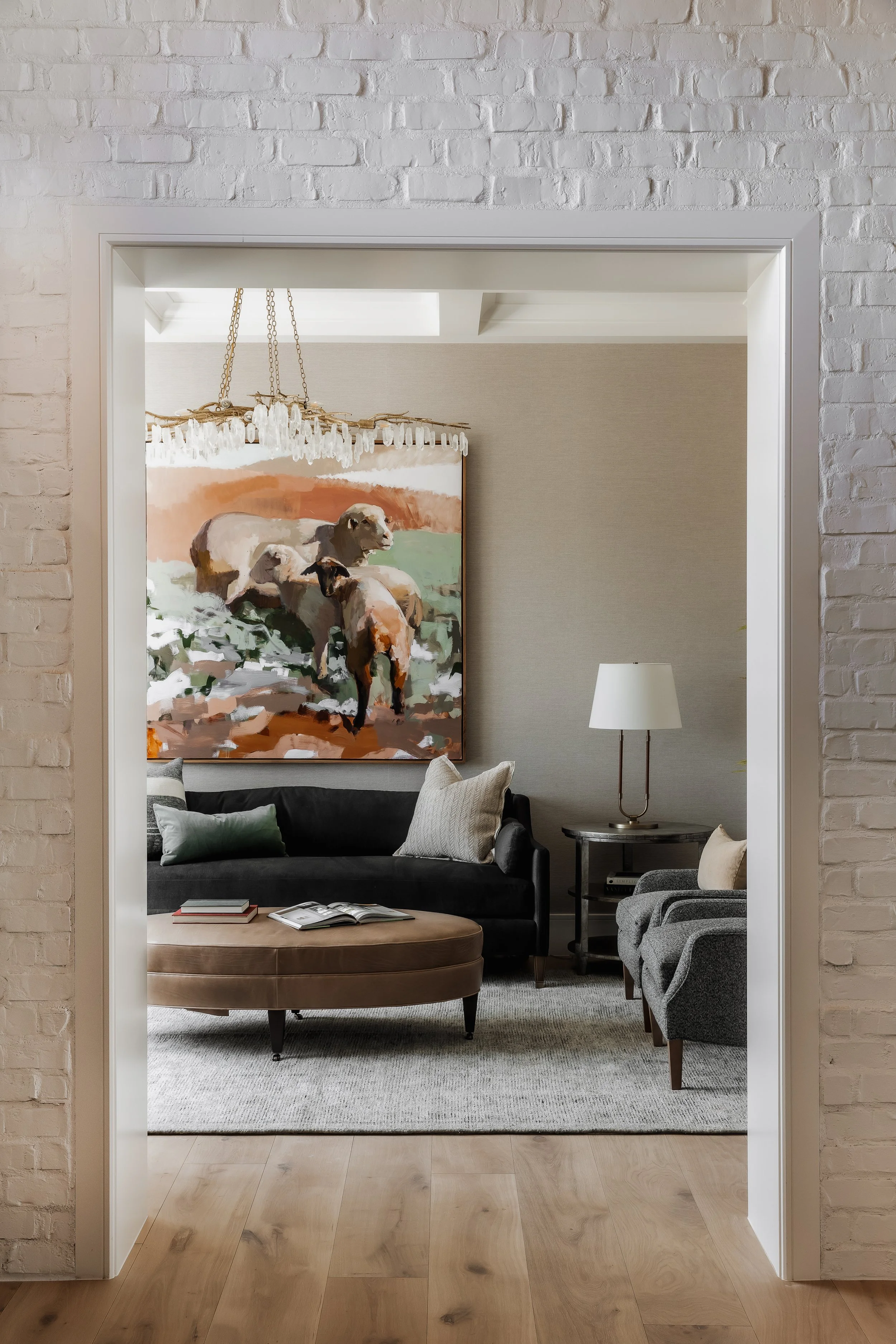 View into a modern living room through a white brick-framed doorway, featuring a black sofa with pillows, a round ottoman with books, a gray armchair, a side table with a lamp, and a large painting of sheep on the wall.